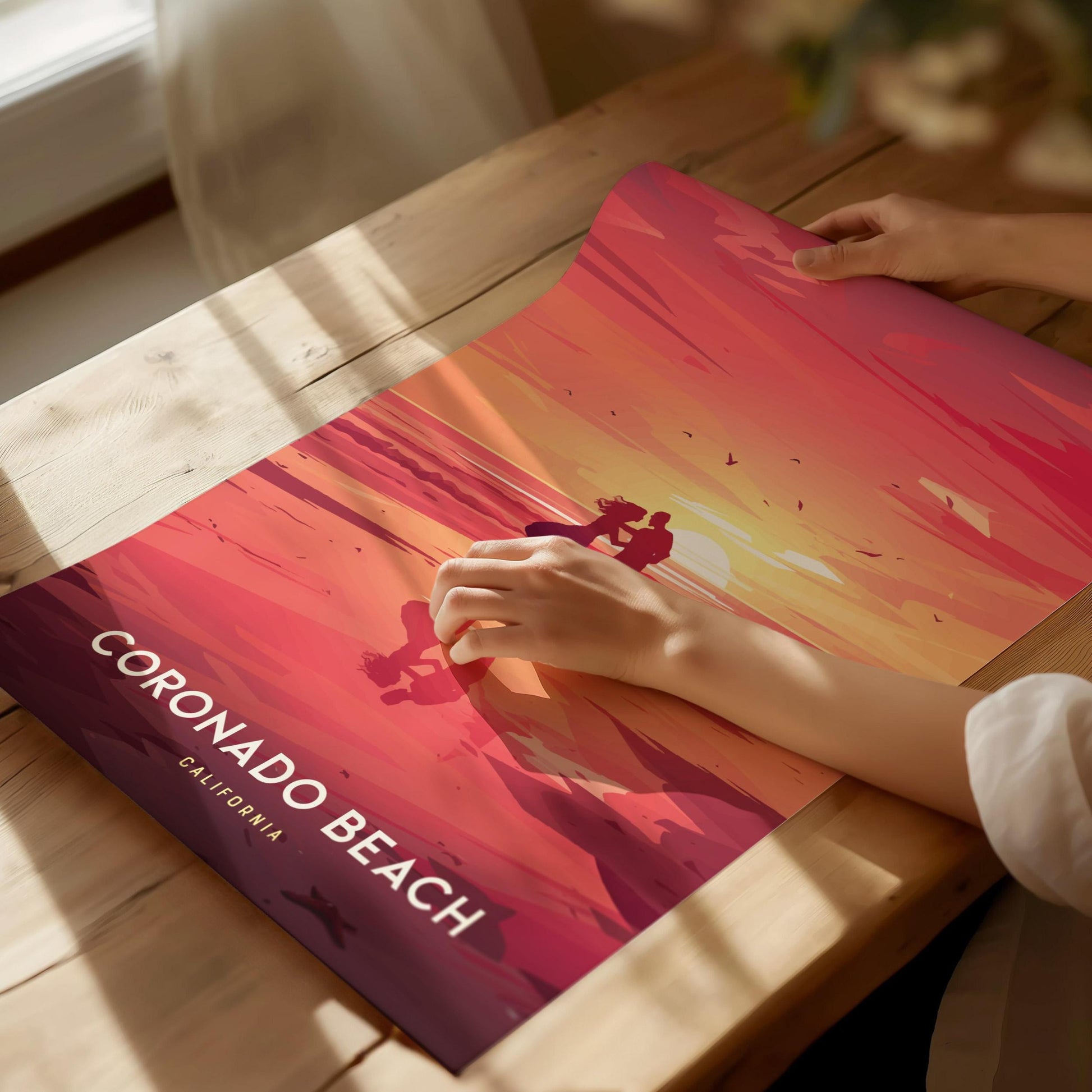 Person holding a poster of Coronado Beach with a sunset design on a wooden table.