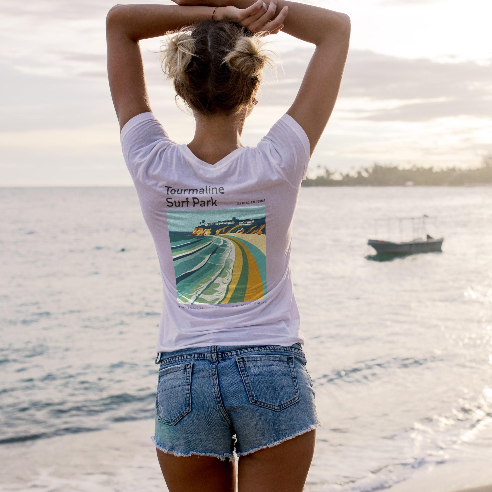 Person wearing a white t-shirt with a graphic design of a surf park and text 'Tourmaline Surf Park' on a beach.