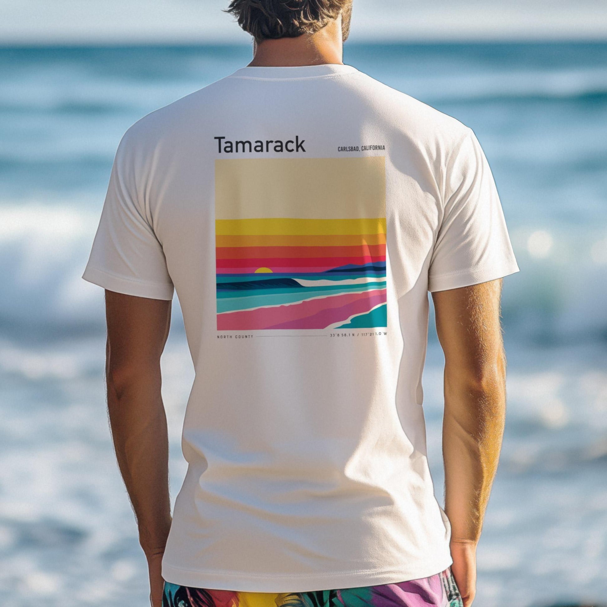 Person wearing a white t-shirt with colorful design and 'Tamarack surf spot' branding at the beach.