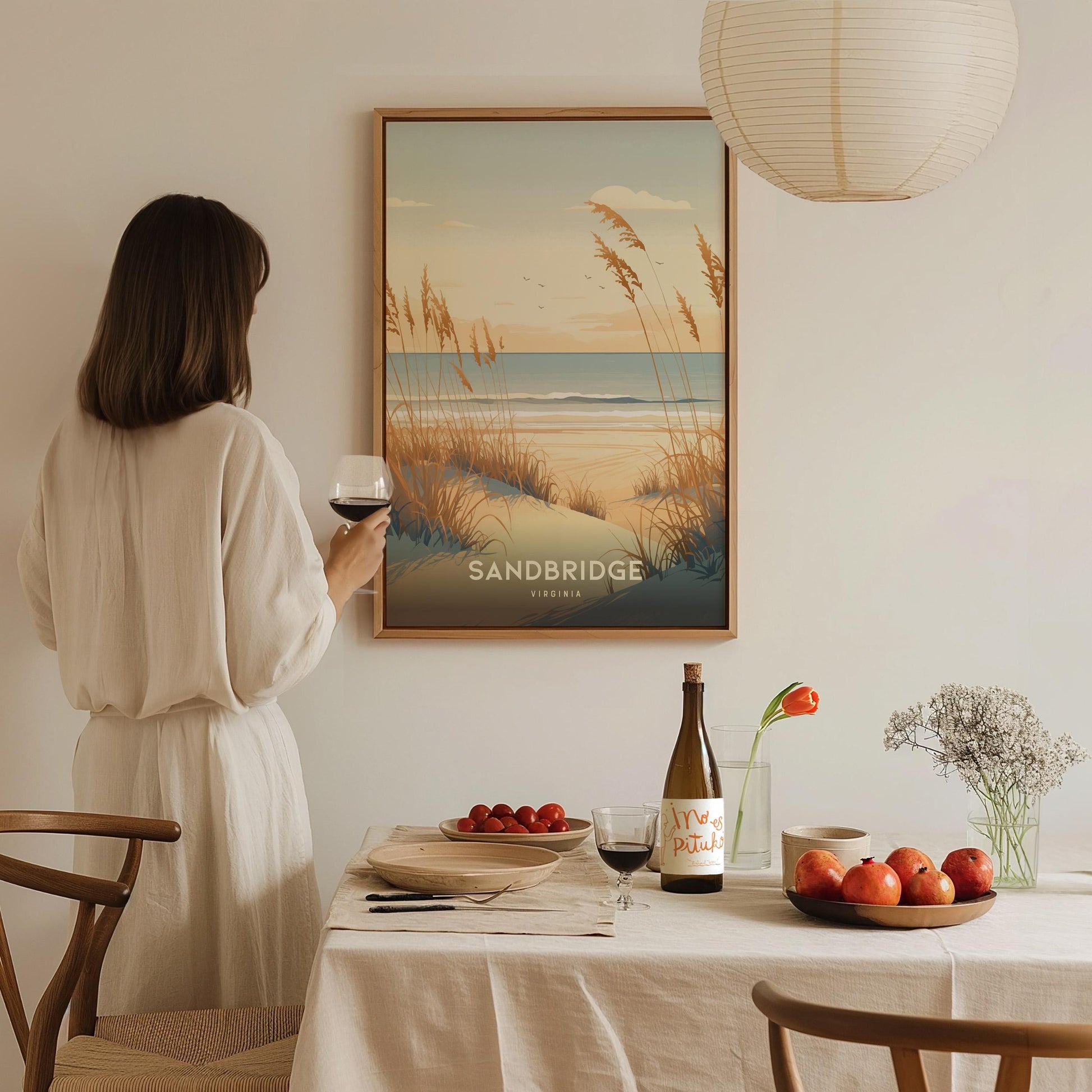 Woman in a white dress standing in a dining room with a 'Sandbridge' beach scene poster on the wall.