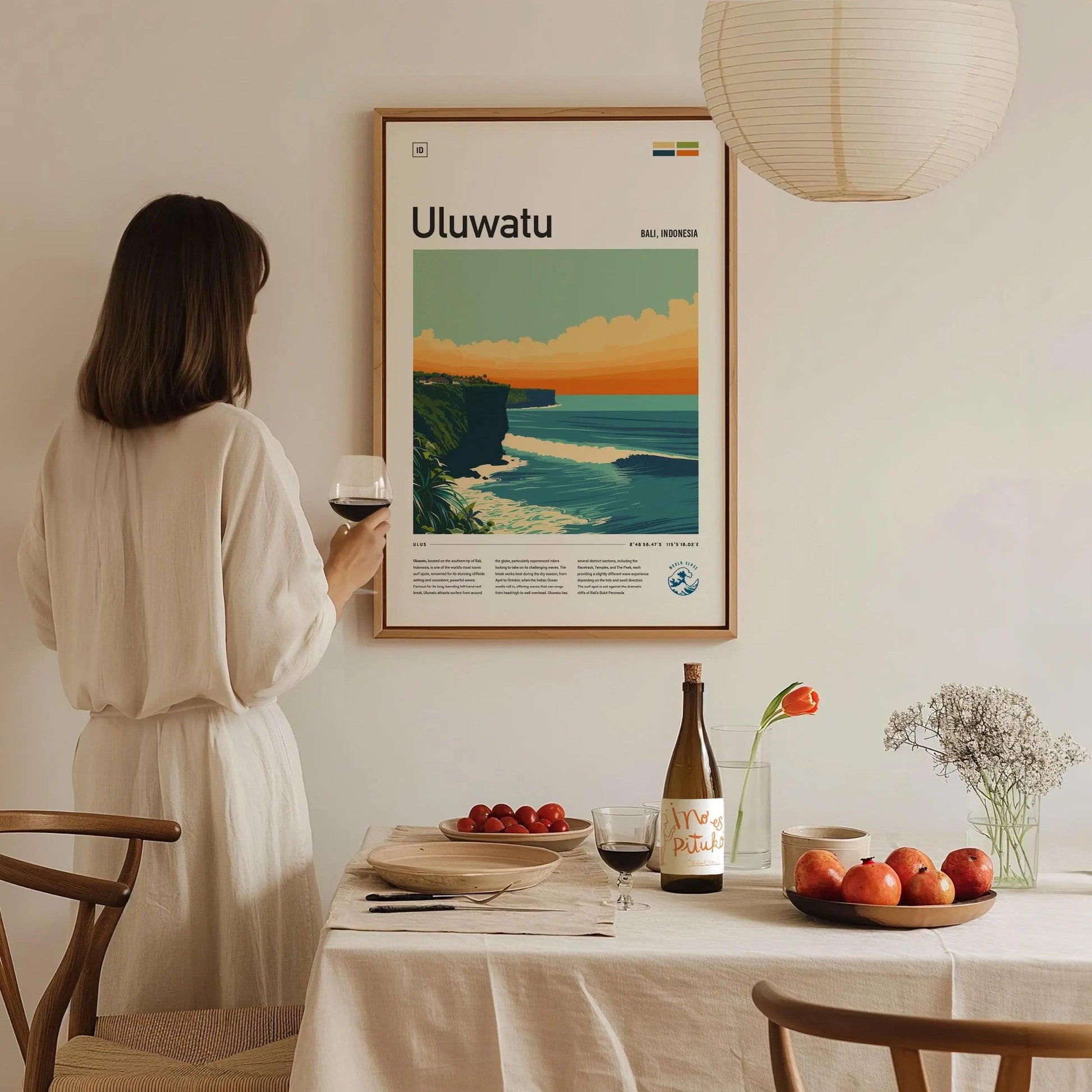 Woman in a white dress standing in a dining room with a framed poster of Uluwatu on the wall.