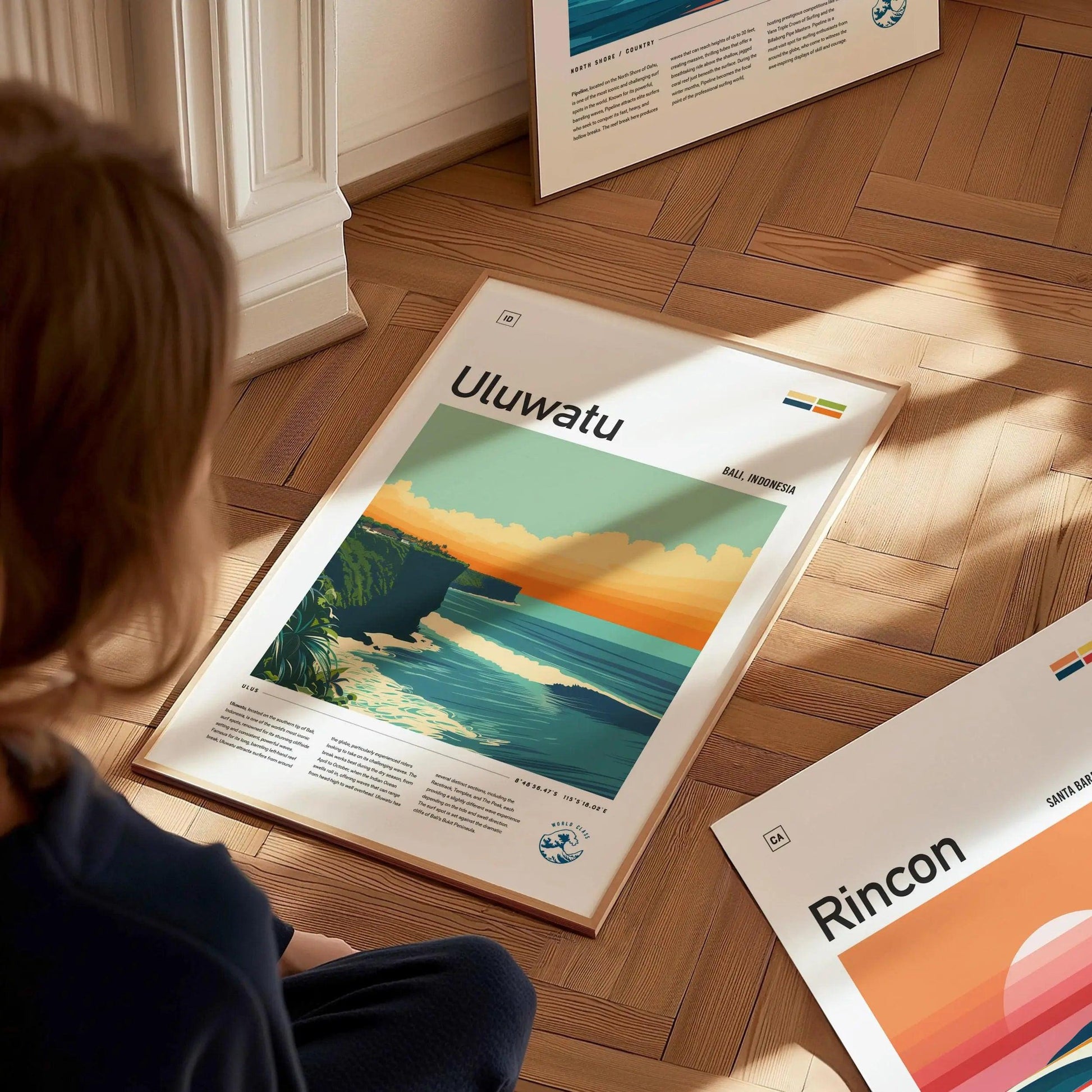 Person looking at a framed poster of Uluwatu on a wooden floor.