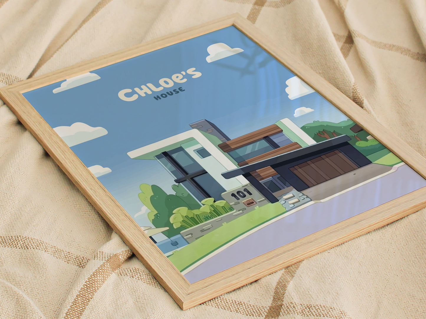 A framed poster of a modern house labeled Chloes House rests on a beige, textured fabric. The design features a bright blue sky with fluffy clouds.