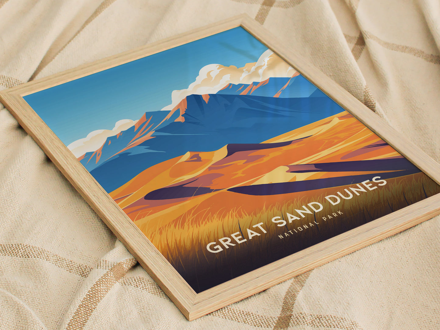A framed poster of Great Sand Dunes National Park rests on a beige fabric, depicting golden dunes against blue mountains and sky.
