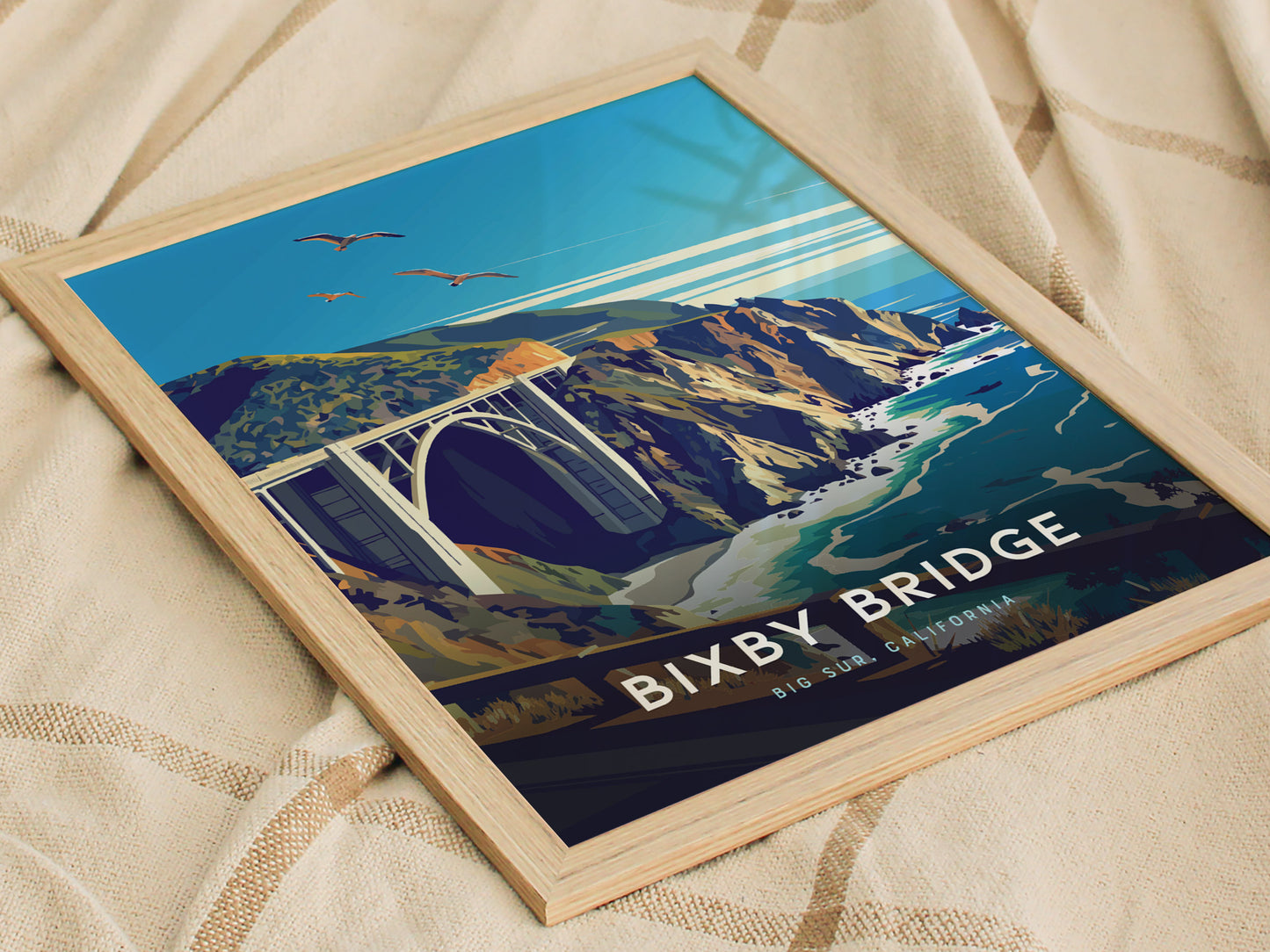 A framed poster depicts the Bixby Bridge in Big Sur, California, with birds flying over rugged cliffs and turquoise waters. The poster is placed on a beige fabric surface.
