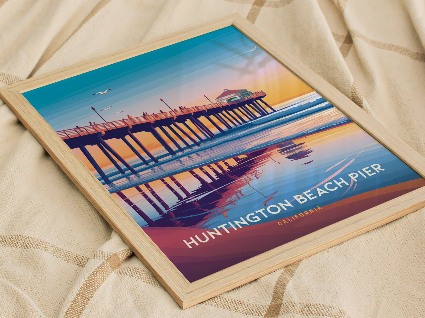 Framed poster of Huntington Beach Pier showcasing a sunset scene with vibrant colors and reflections on the water. The image is placed on a textured beige fabric.