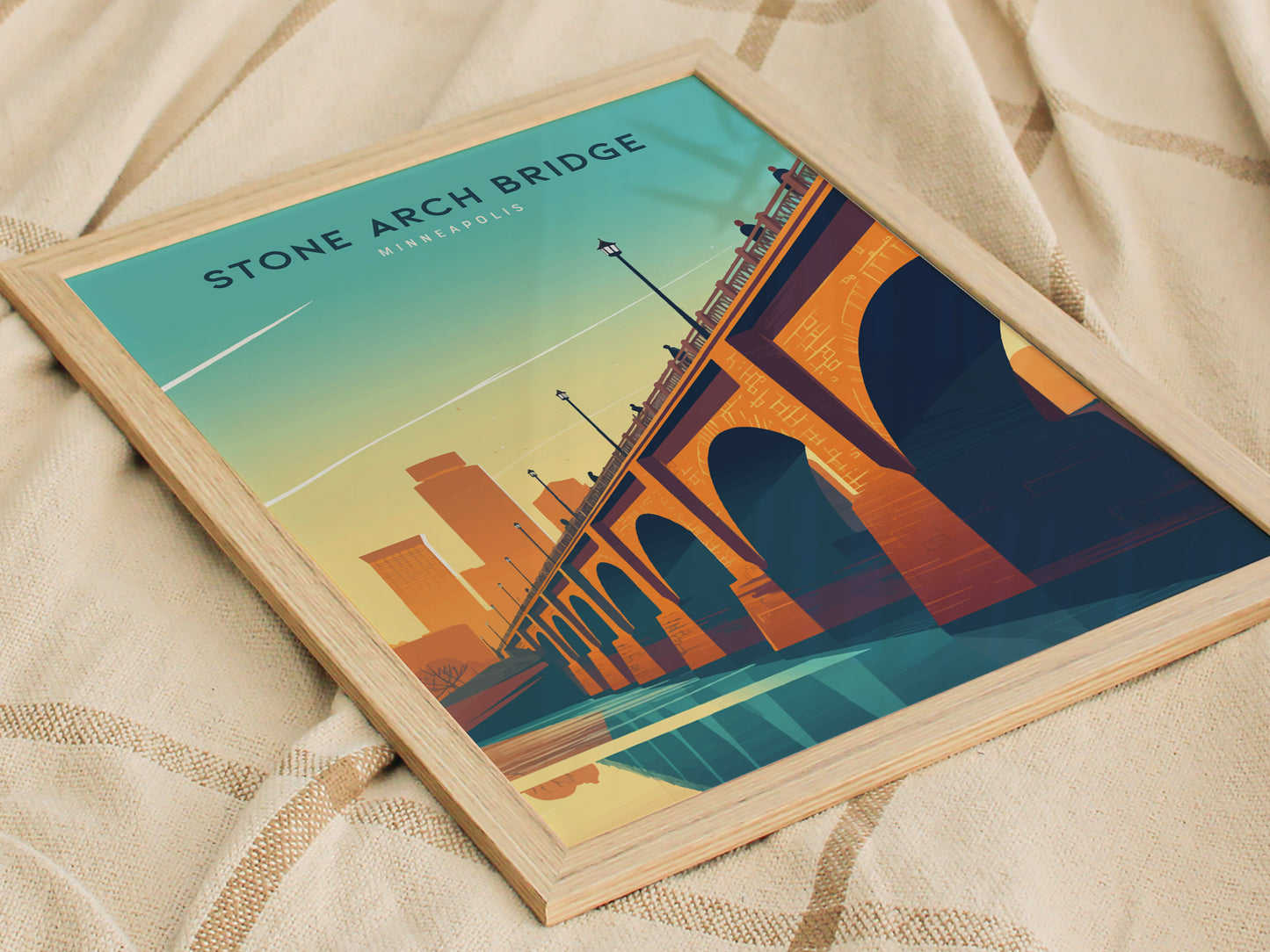 A framed poster of the Stone Arch Bridge with a stylized Minneapolis skyline is placed on a beige textured blanket. The artwork features vibrant orange.