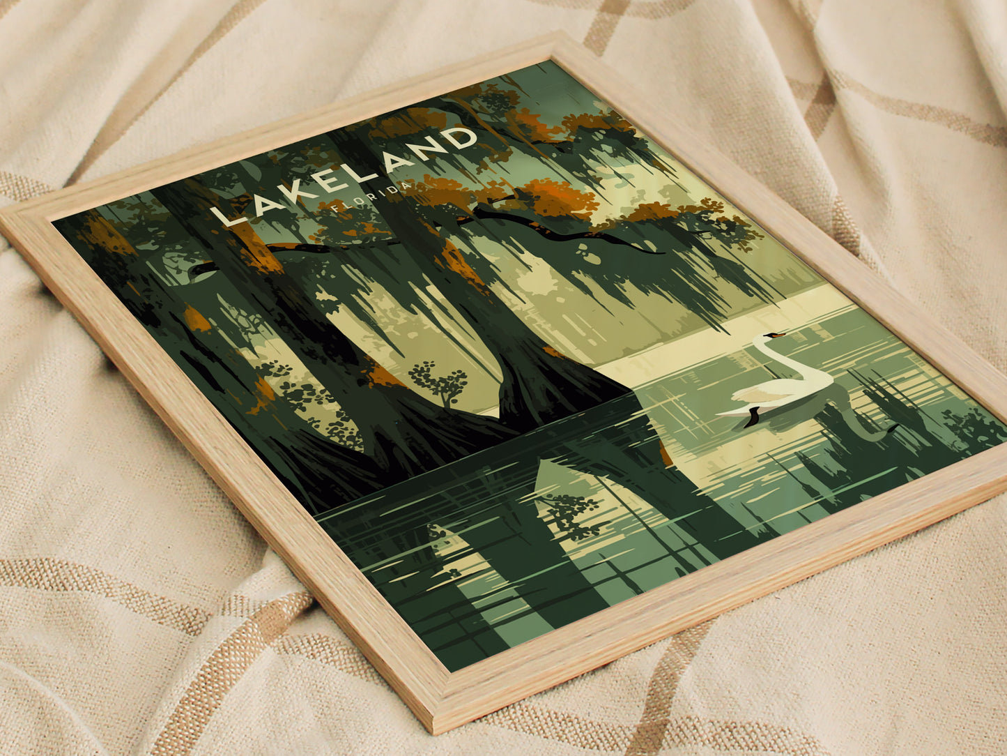 A framed poster of Lakeland, Florida, depicting a serene lake scene with cypress trees and a swan, rests on a textured fabric surface. The artwork uses earthy, muted colors.