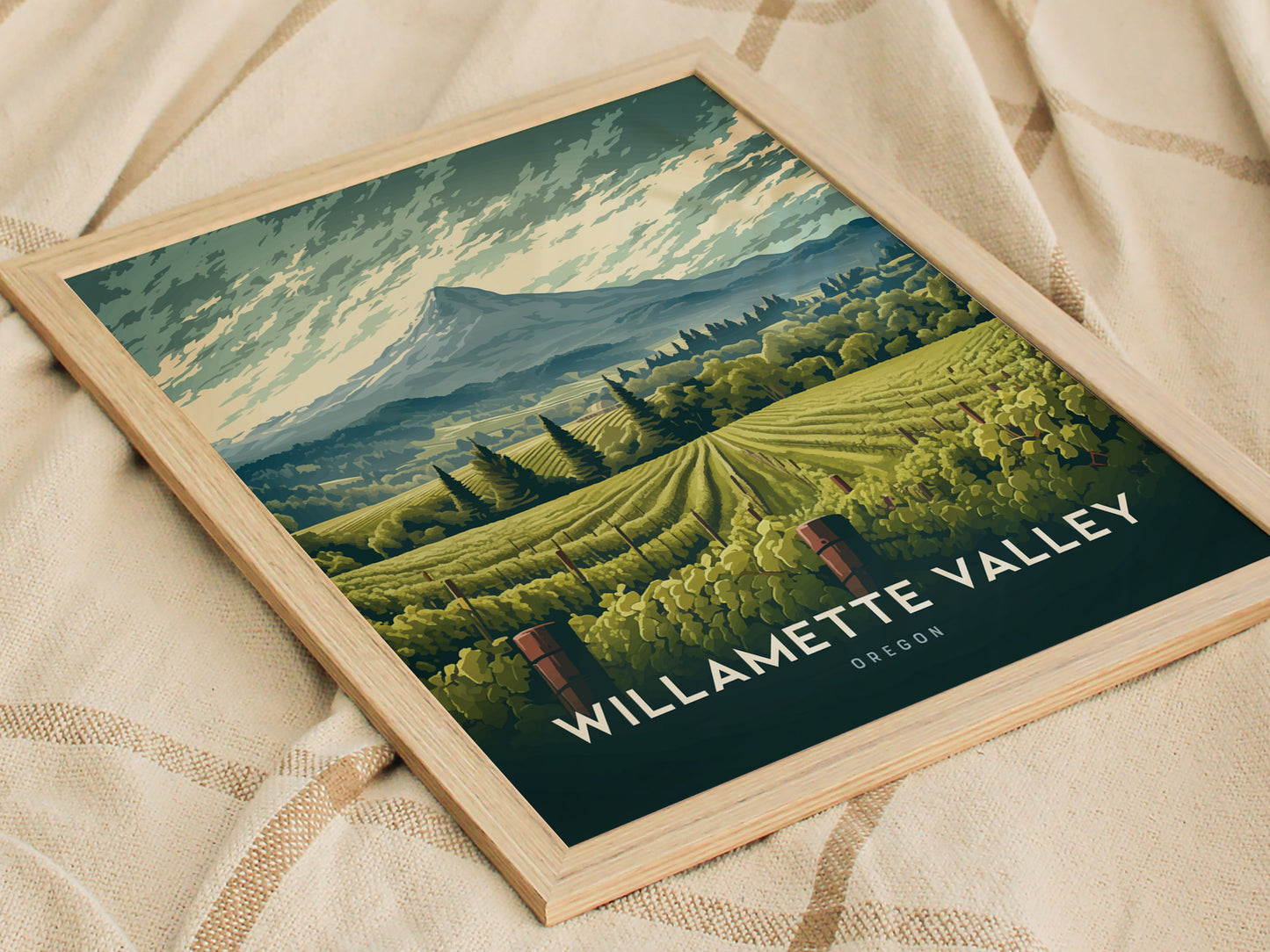 A framed illustration of Willamette Valley, Oregon, featuring rolling vineyard hills and a mountainous backdrop, rests on a textured fabric surface. The artwork highlights lush greenery and serene landscapes.