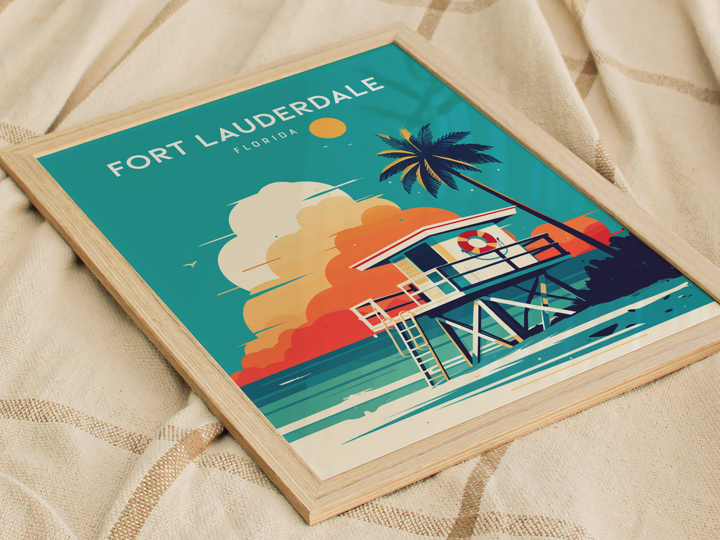 A framed poster depicting a vibrant illustration of Fort Lauderdale Beach, featuring a lifeguard stand, a coconut palm, and a colorful sky, resting on a textured fabric surface.
