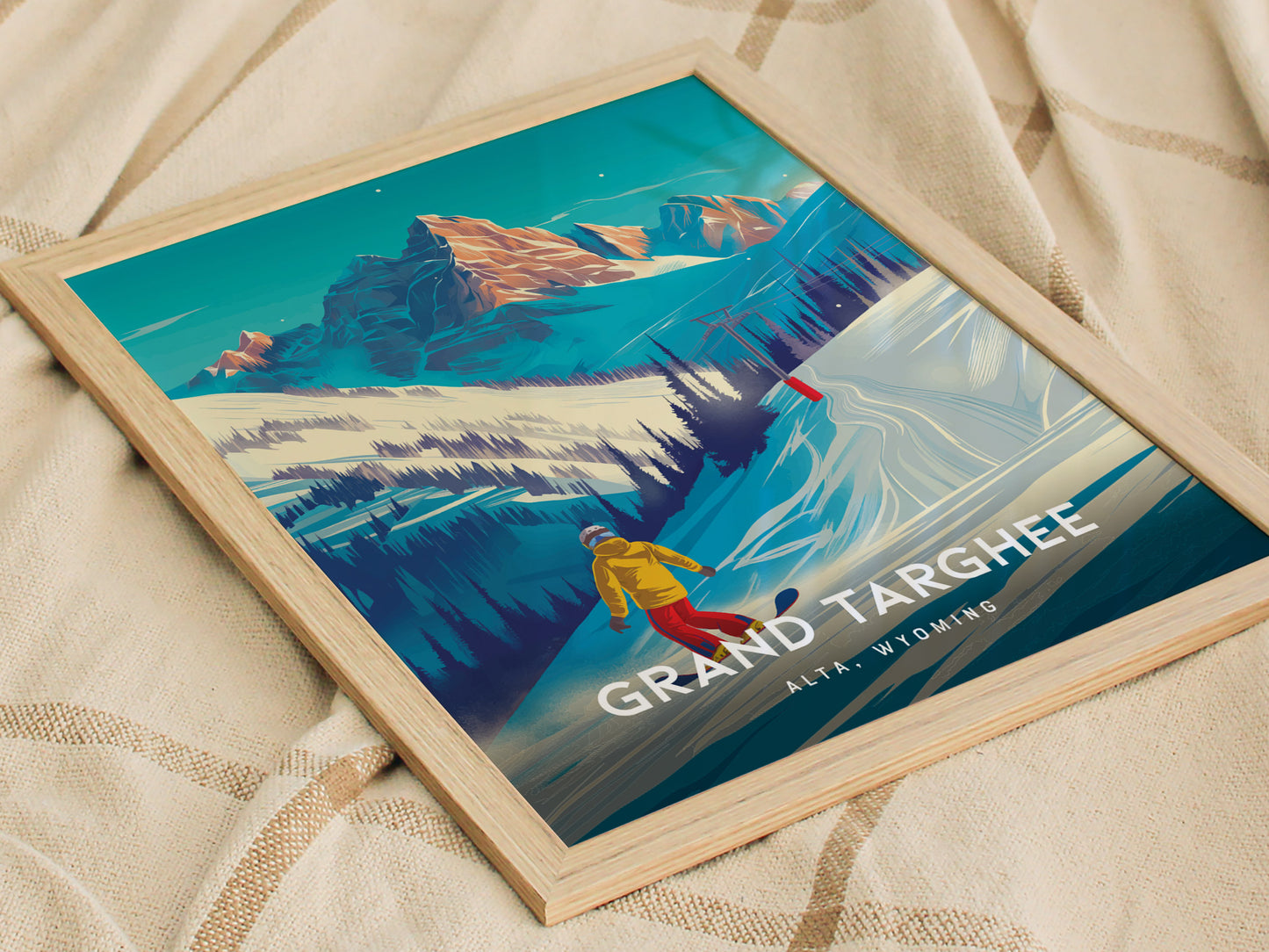 A framed poster featuring a snowboarder in a yellow jacket on a snowy slope, with the Grand Targhee Resort and Teton Mountains in the background, placed on a beige fabric.