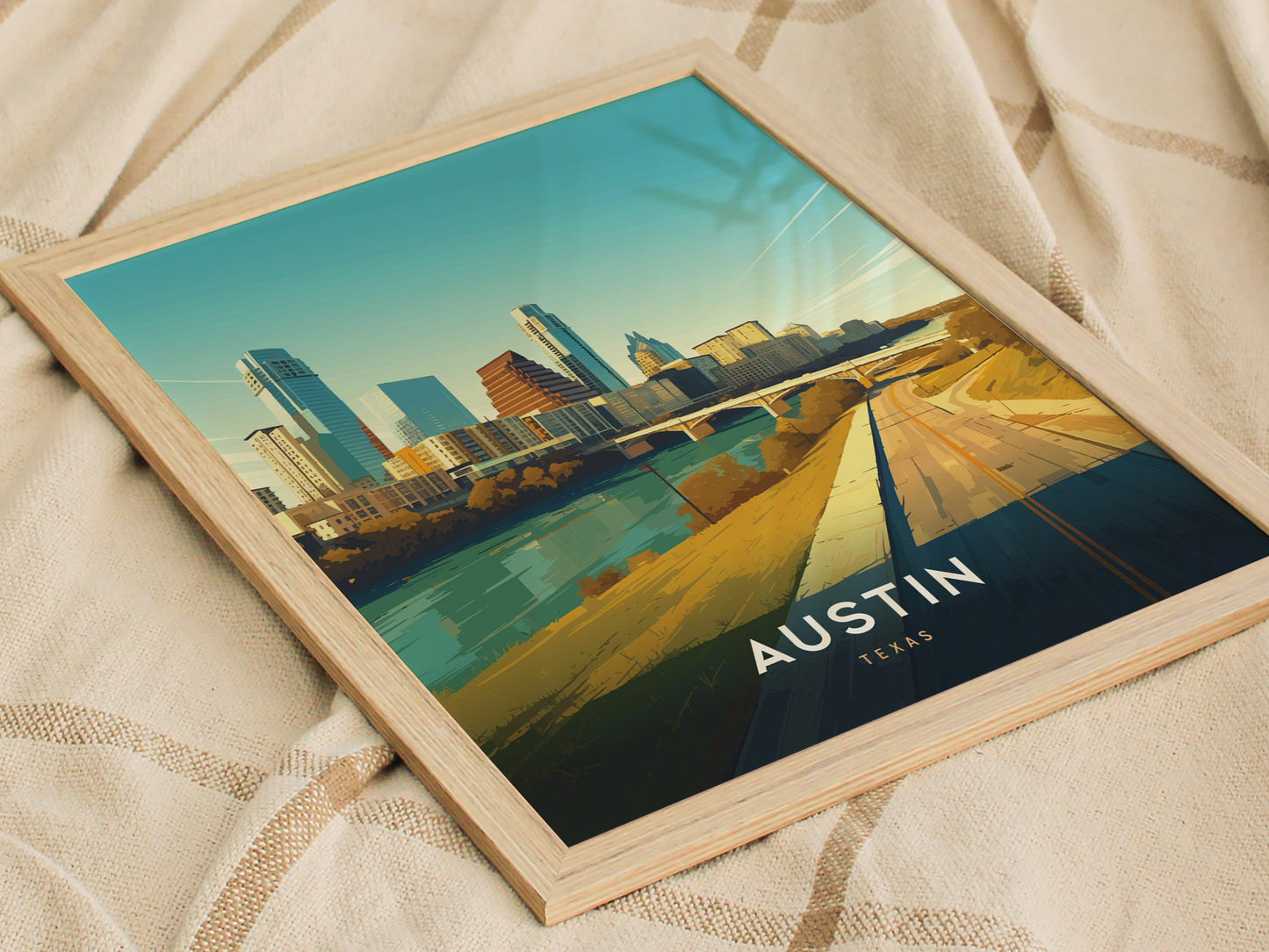 Framed artwork depicting Austin, Texas skyline with modern buildings and a serene river, placed on a textured fabric surface. The vibrant colors highlight the citys dynamic architecture.
