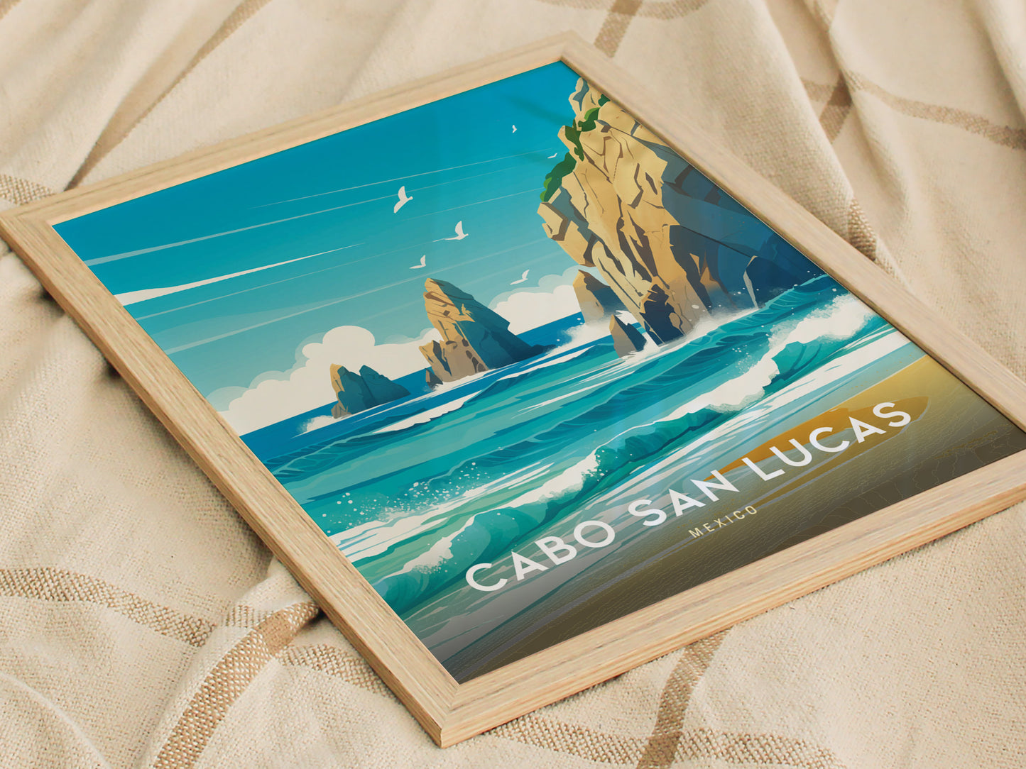 Framed poster of Cabo San Lucas displaying vibrant blue waters, dramatic rock formations, and seagulls in flight, placed on a textured beige fabric background.