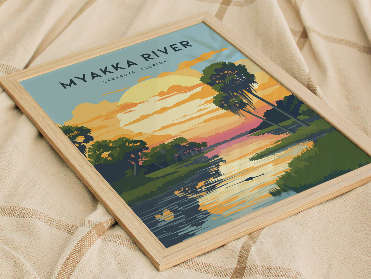 Framed Myakka River State Park poster resting on a textured beige fabric, depicting a vibrant sunset with palm trees and a reflective river scene in Sarasota, Florida.