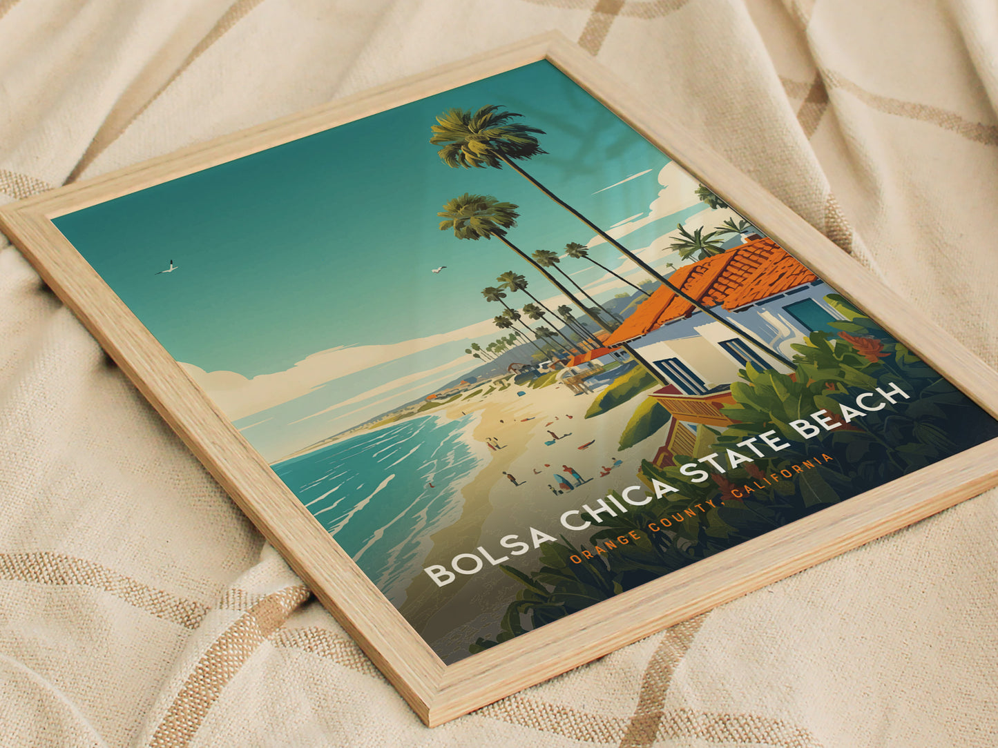 A framed Bolsa Chica State Beach poster features a coastal scene with palm trees, sandy beach, and ocean waves under a blue sky, set on a beige fabric background.