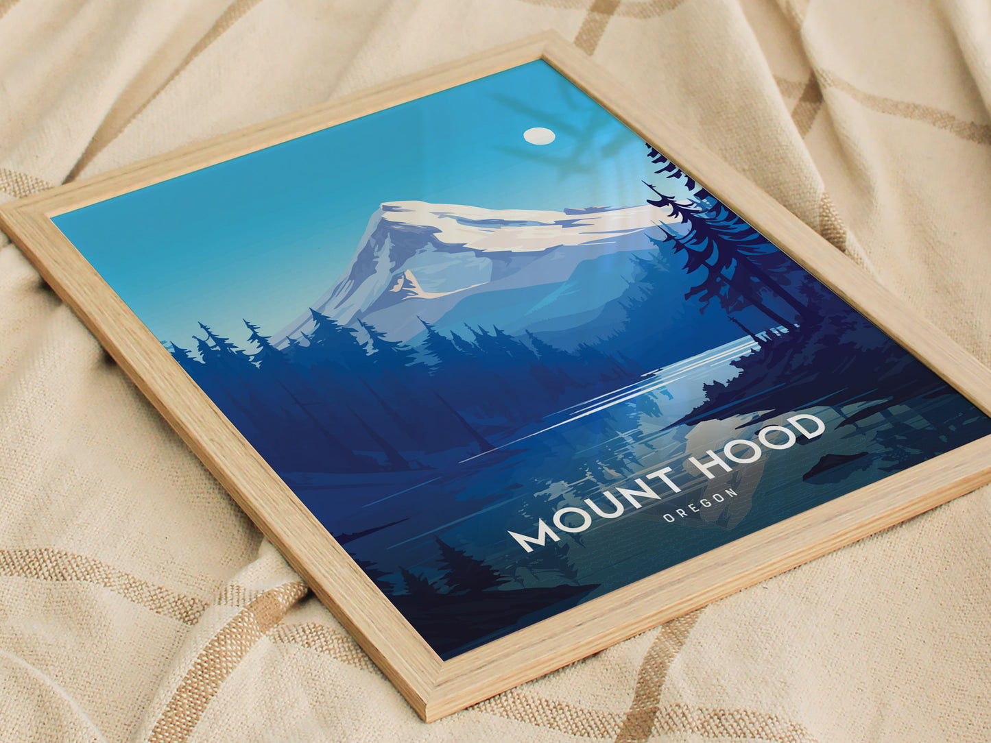 Framed poster of Mount Hood featuring a stylized illustration with snow-capped peaks, forested foothills, and a clear blue sky, resting on a textured beige fabric.