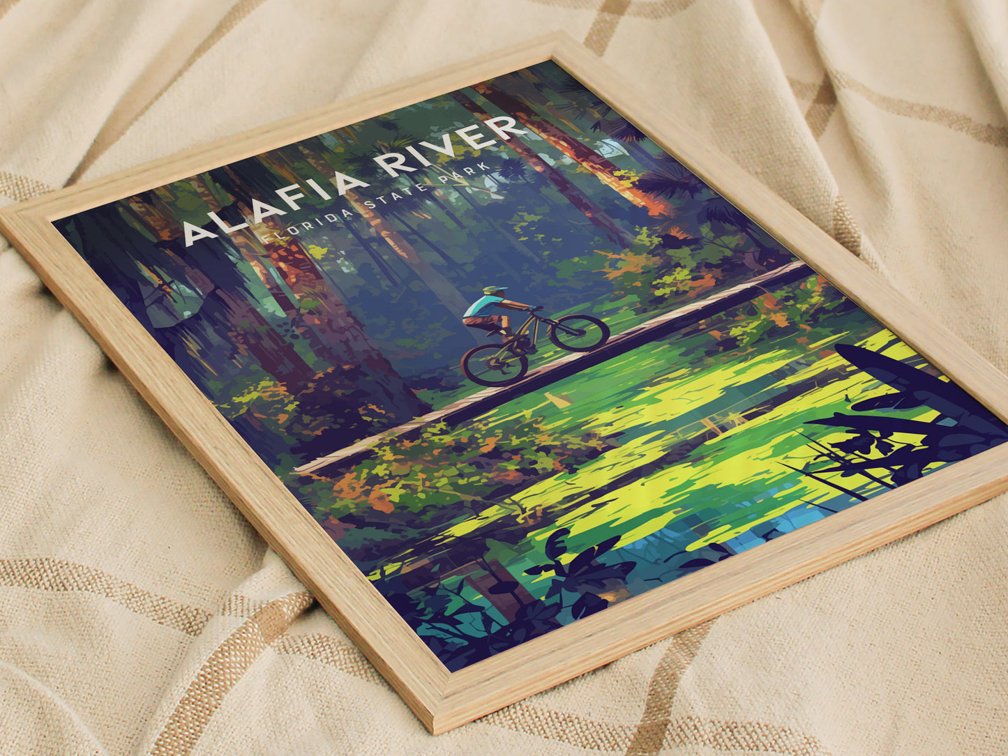 Framed poster of a cyclist on a wooden bridge in a vibrant forest scene, captioned Alafia River Florida State Park, resting on a textured beige fabric.