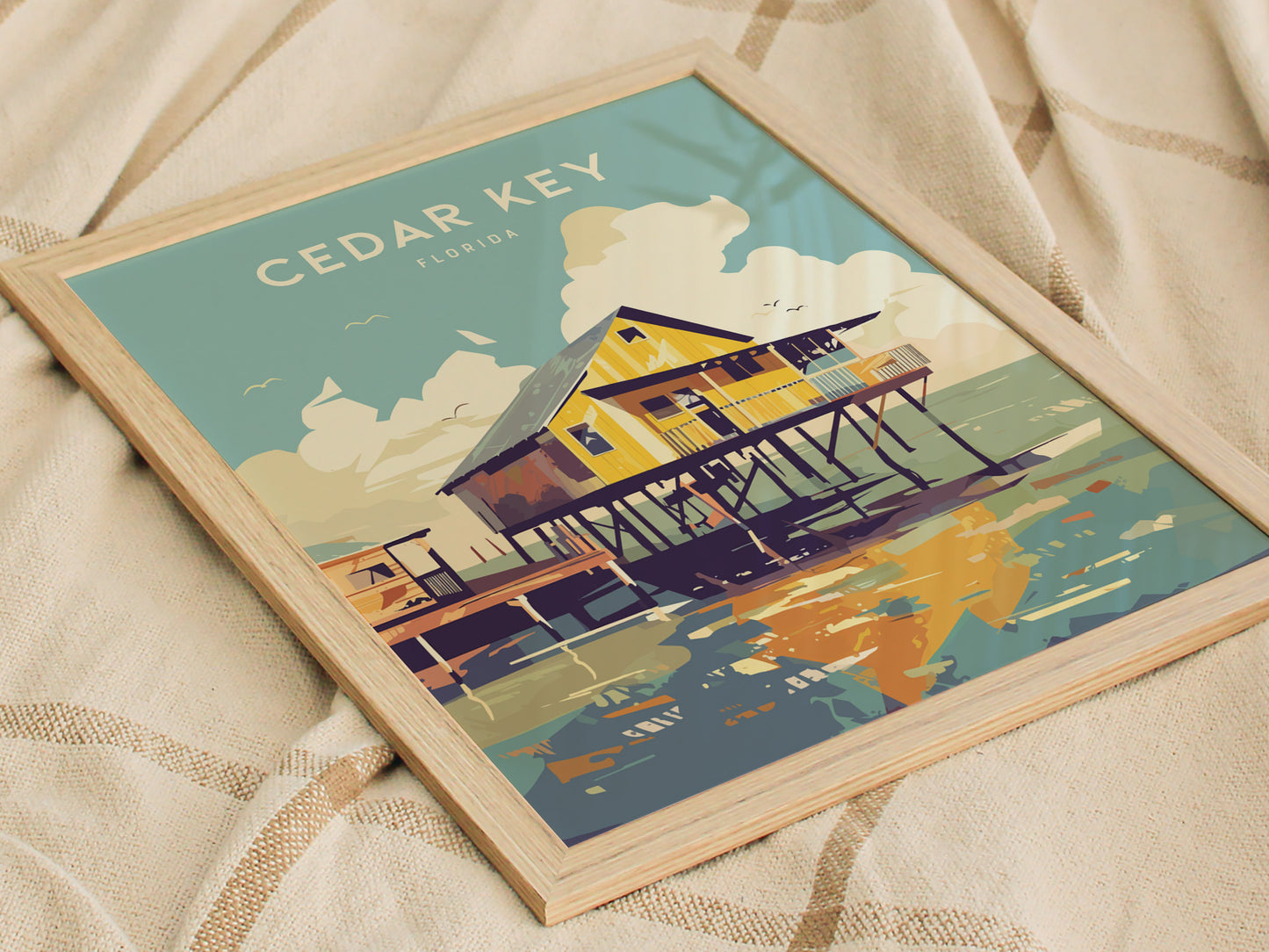 Framed poster of a stilt house over water, with Cedar Key Florida text, placed on a textured beige fabric. The artwork features vibrant colors and a serene coastal scene.