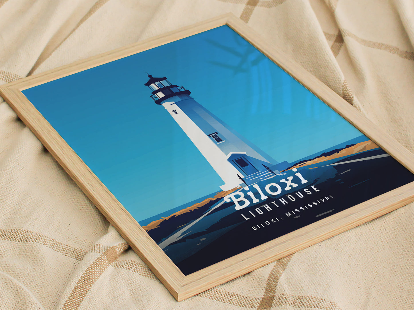 Framed artwork of Biloxi Lighthouse in Mississippi, featuring a bold, minimal vector design with a bright blue sky, placed on a patterned fabric surface.