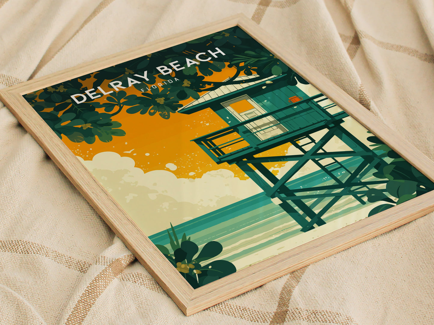 Framed artwork of a lifeguard stand against an orange sky with sea grape leaves and ocean waves, labeled Delray Beach Florida, resting on a beige fabric background.