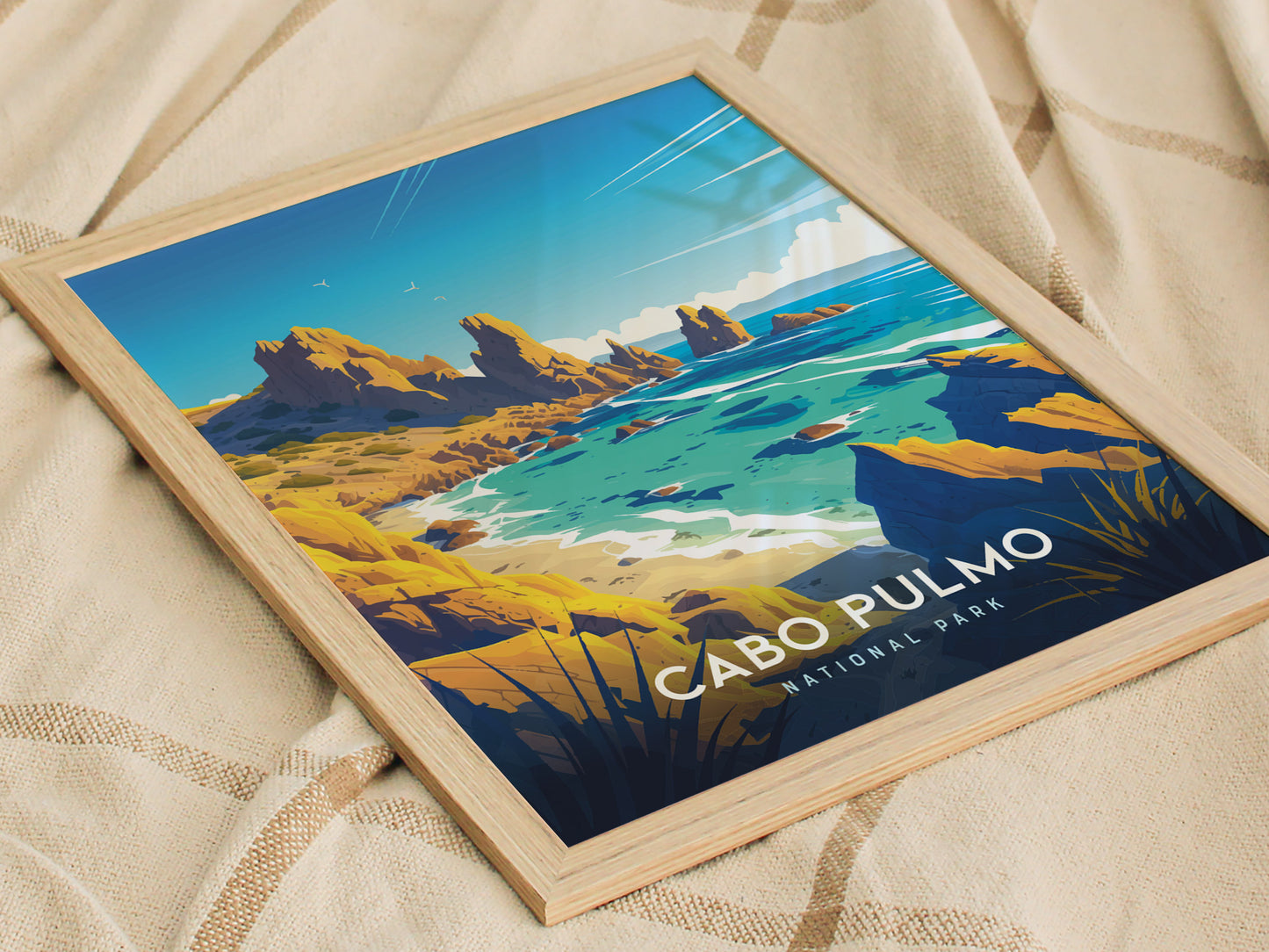 A framed art print of Cabo Pulmo National Park features a vibrant coastal landscape with rocky cliffs, turquoise waters, and seagulls, resting on a beige fabric background.
