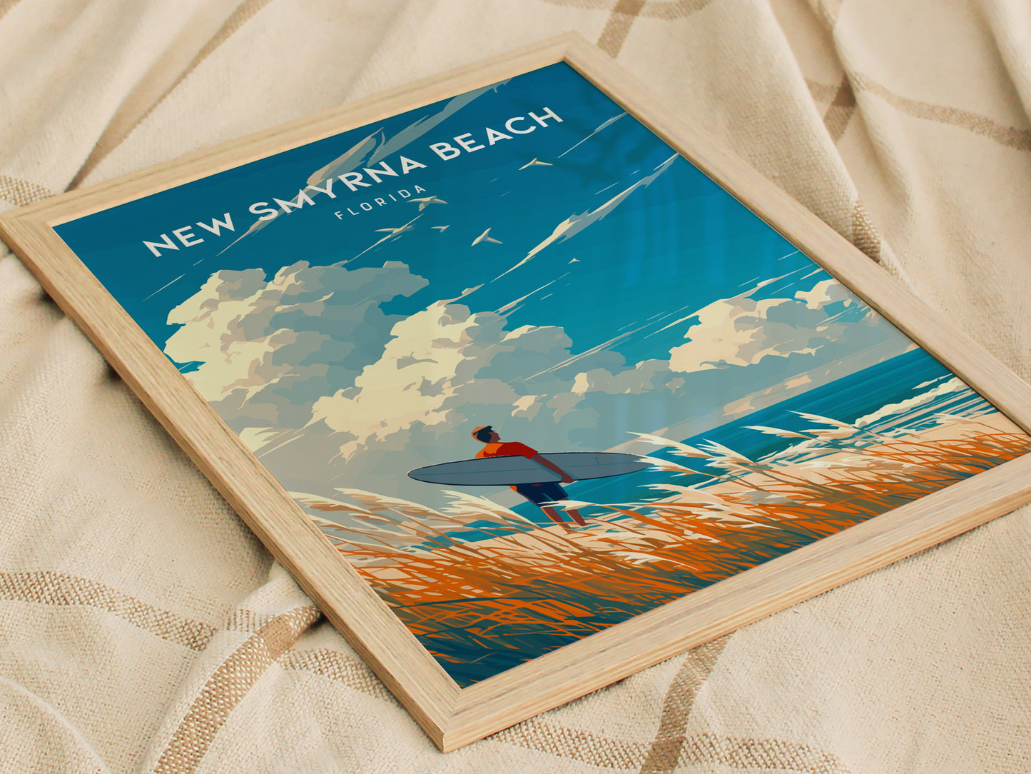 Framed poster featuring a surfer silhouette carrying a surfboard against a coastal backdrop with clouds and waves, labeled New Smyrna Beach, Florida. The poster rests on a textured fabric.
