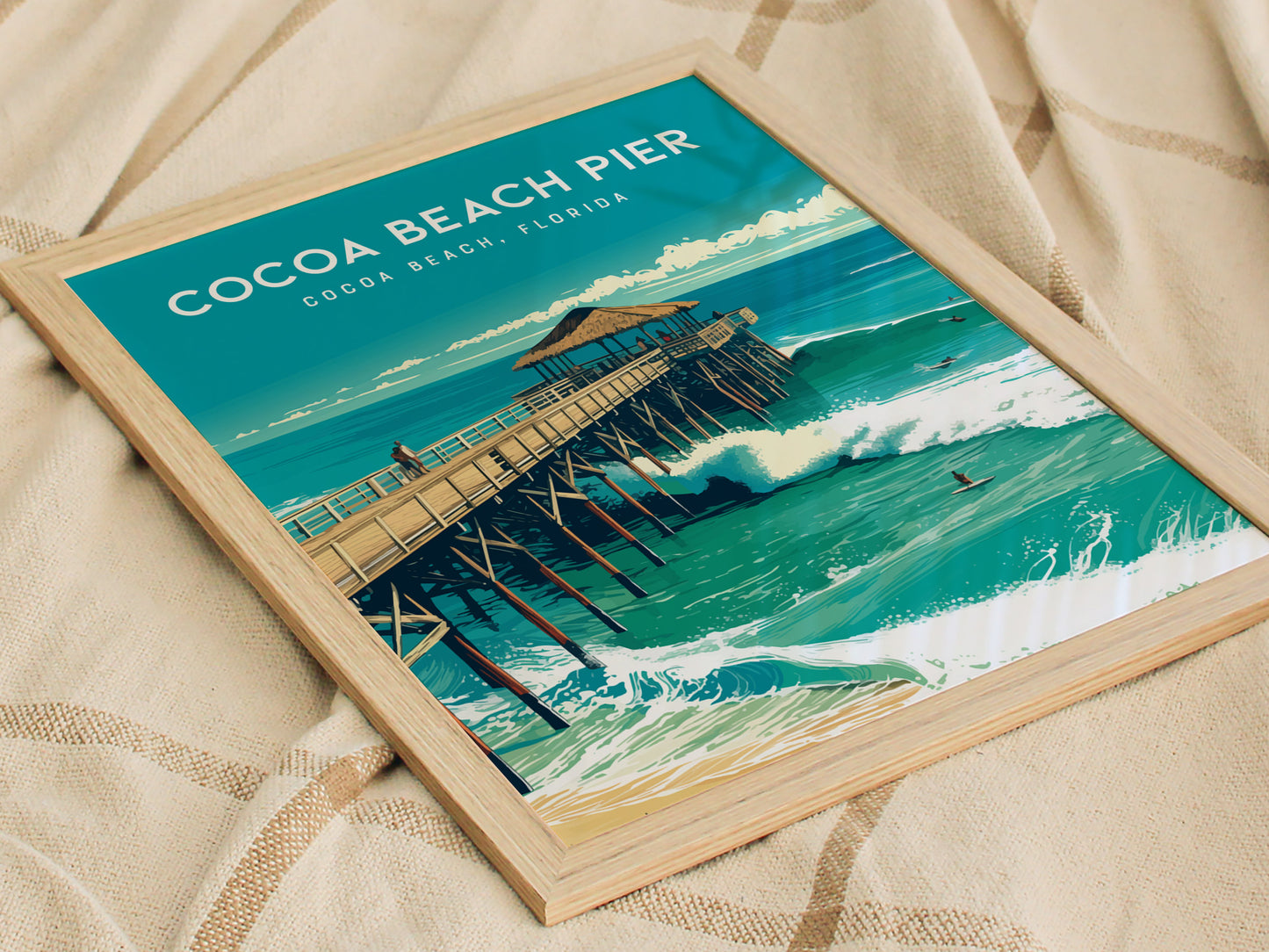 Framed artwork depicts Cocoa Beach Pier extending over vibrant, turquoise waves with a thatched-roof pavilion. Surfers navigate the ocean below, capturing the essence of Floridas coastal charm.