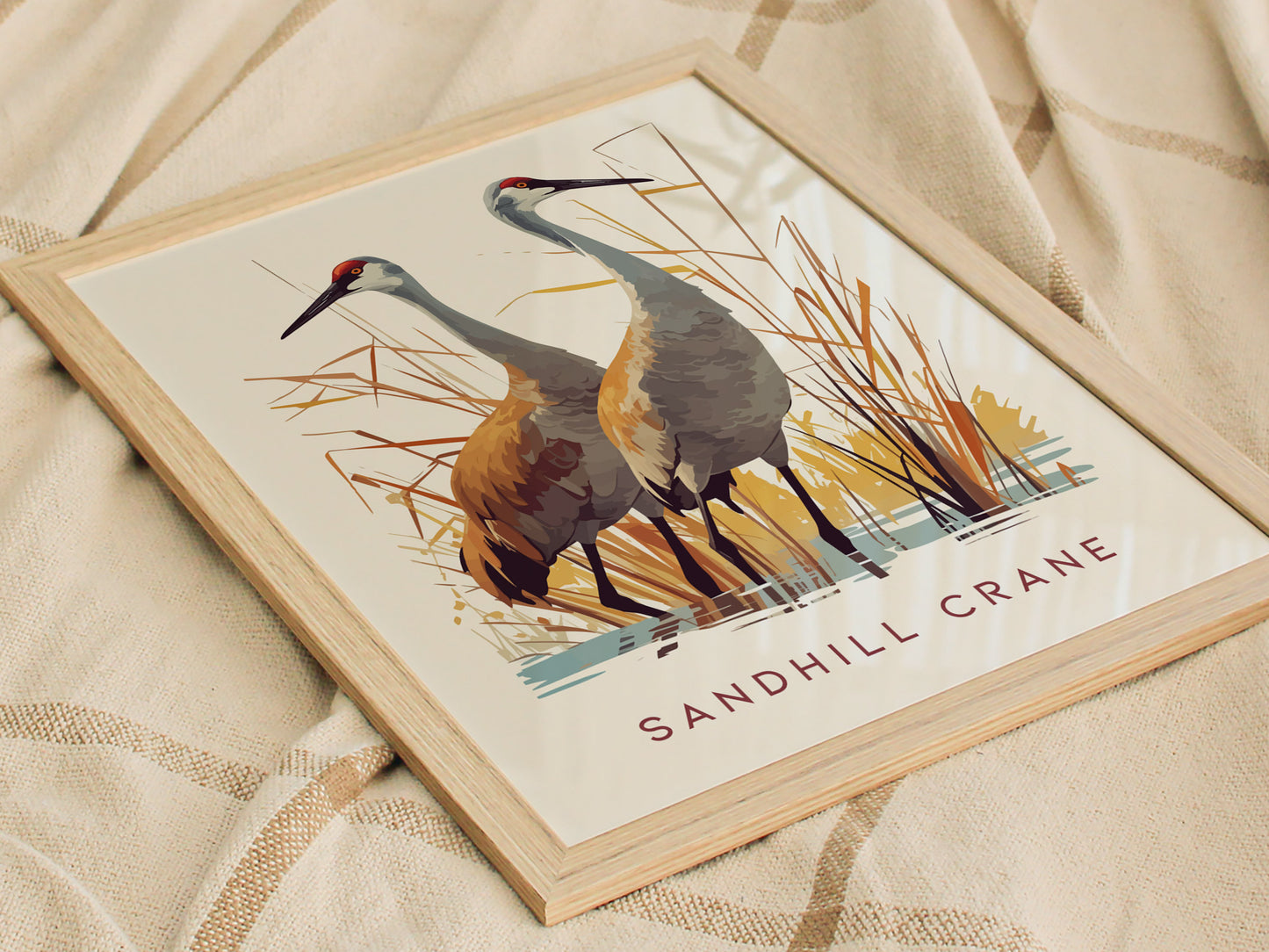 A framed poster displays two sandhill cranes standing amidst stylized marsh grasses with the text SANDHILL CRANE beneath, resting on a textured beige fabric.