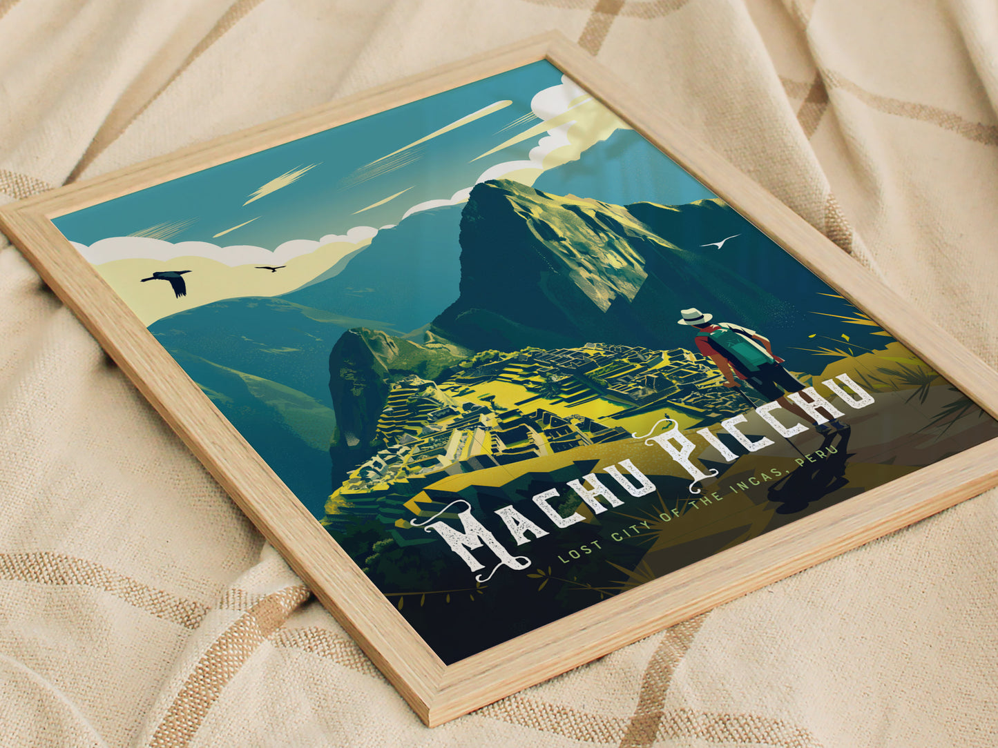 Framed artwork depicting Machu Picchu with stylized mountains and a hiker, set against a sky with birds. The image rests on a textured beige fabric.