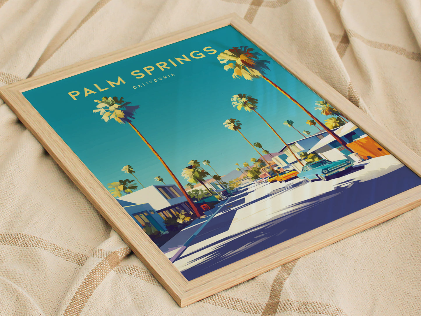 A framed poster of Palm Springs, California, features palm trees, mid-century architecture, and vintage cars under a clear blue sky, resting on a textured fabric.