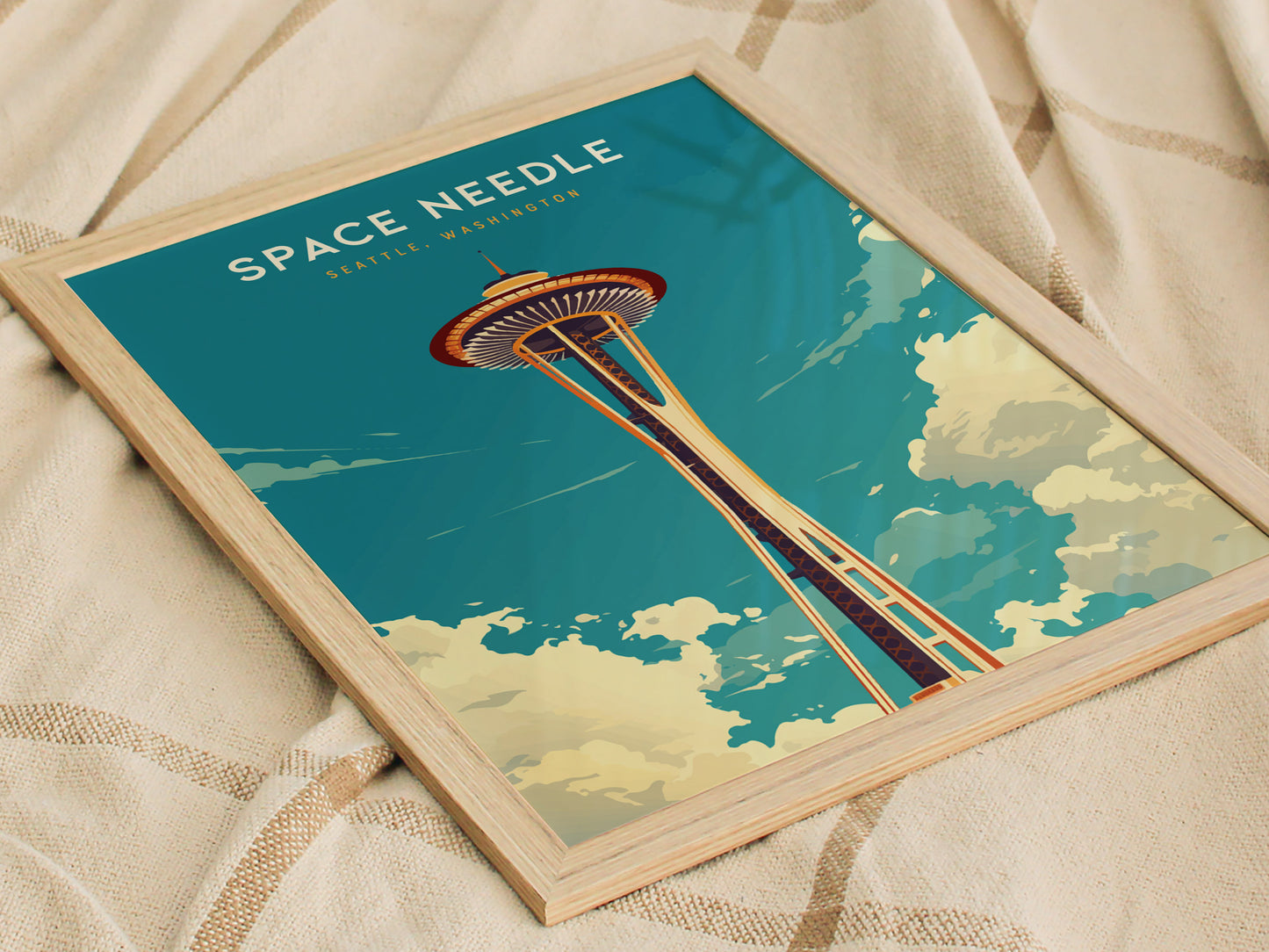 Framed poster of the Space Needle against a blue sky with clouds, laid on a beige fabric. The text reads Space Needle, Seattle, Washington in white letters.