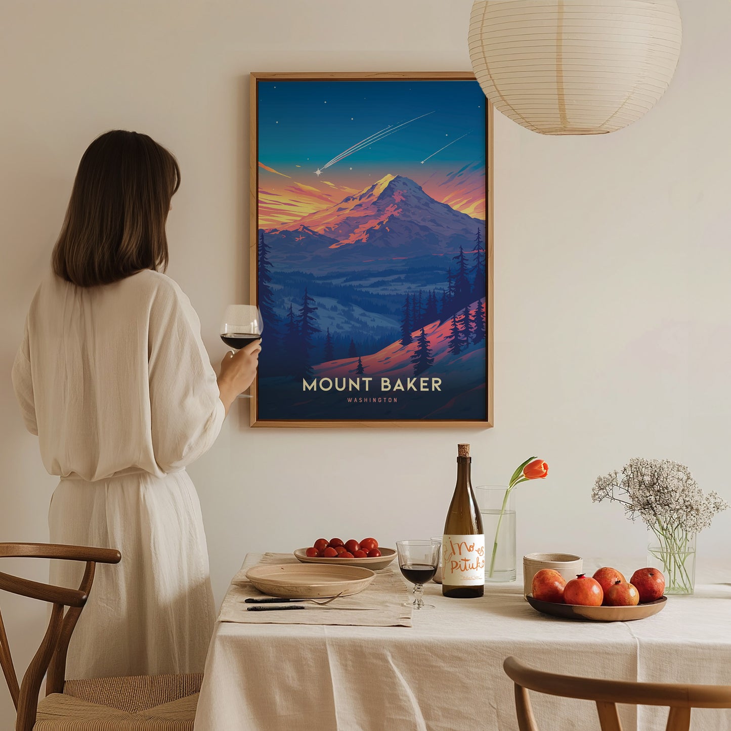 A woman in a white dress holds a glass of wine, admiring a colorful Mount Baker poster in a dining room with a table set for a meal.