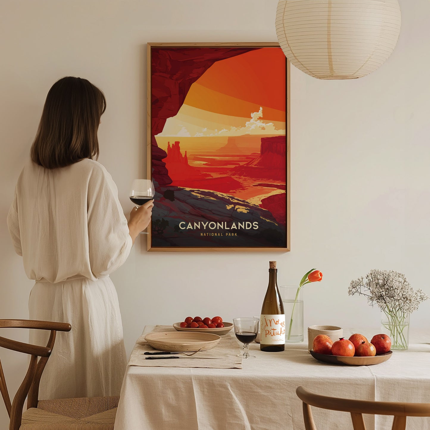 A woman in a white dress admires a framed Canyonlands National Park poster. The table in front holds wine, apples, and tomatoes, creating a cozy, artistic dining setting.