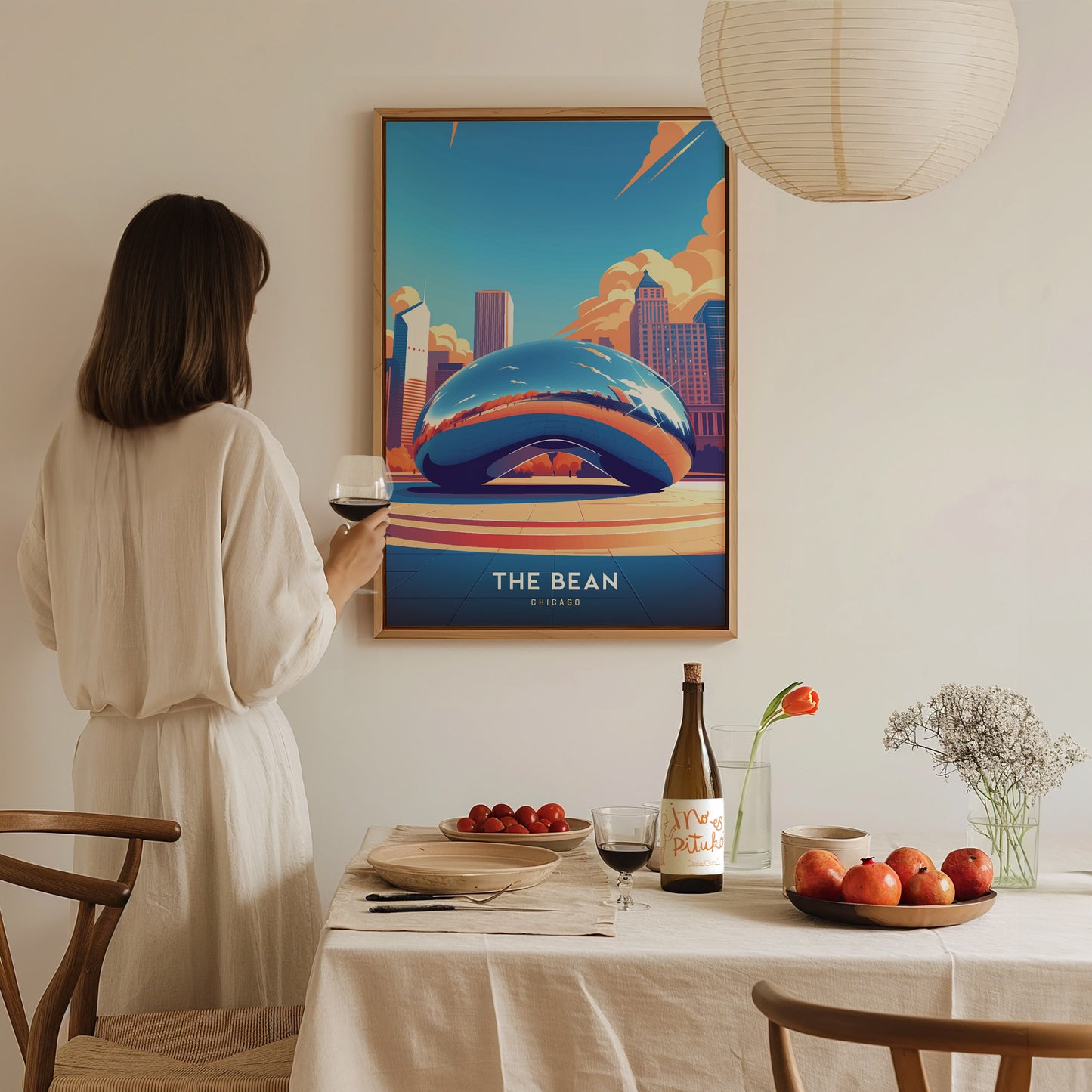 A person in a white dress holds a glass of wine, admiring a framed poster of The Bean with a dining table set with fruit and flowers.