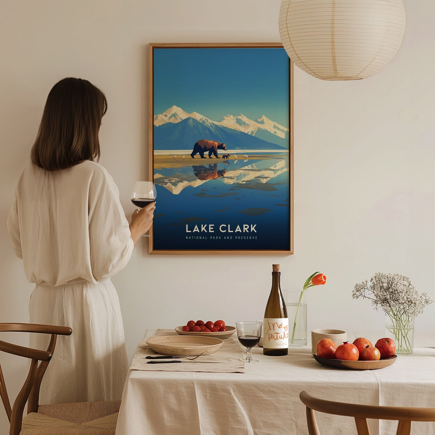 A woman in a white dress holds a glass of wine, looking at a framed Lake Clark National Park poster. A dining table with fruit and wine is in the.