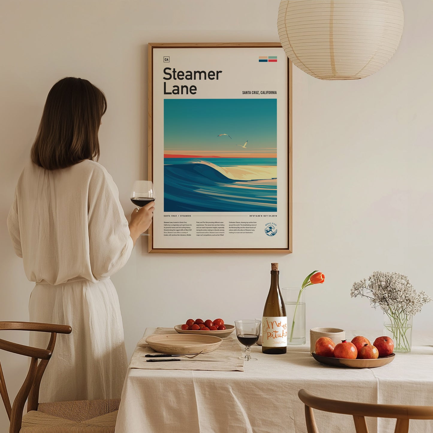 A woman in a white dress holds a wine glass, admiring a Steamer Lane poster on the wall. A table with fruit, a wine bottle, and flowers is in the.
