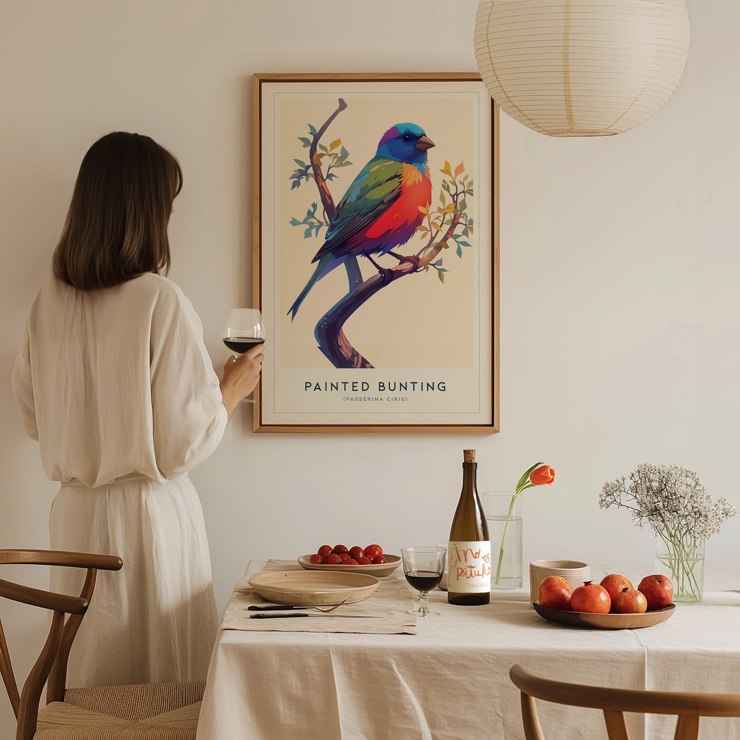A woman in a light-colored outfit stands by a dining table, admiring a colorful art print of a Painted Bunting on the wall. The table is set with food, drinks, and a vase of flowers, creating a cozy and inviting atmosphere.