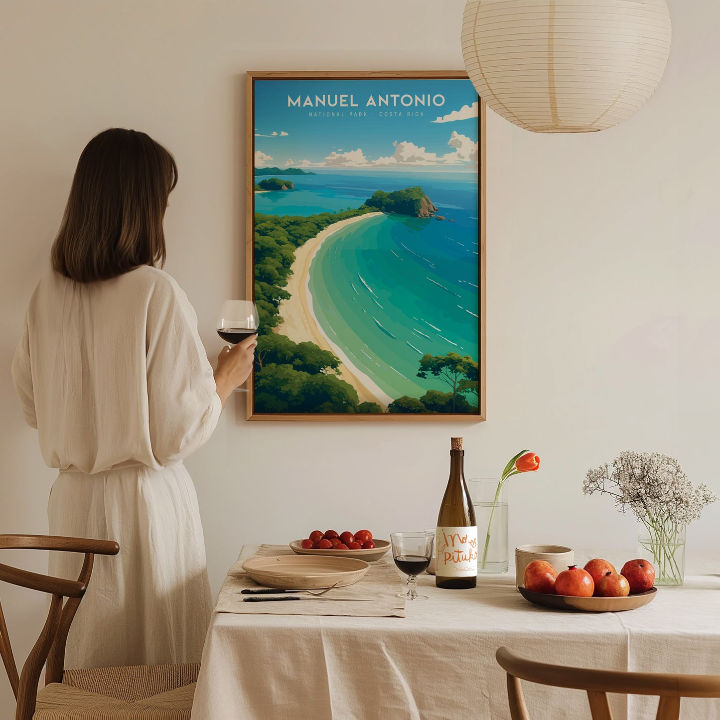 A woman in a white dress holds a glass of wine, admiring a framed Manuel Antonio National Park poster. The table is set with fruit, wine, and flowers.