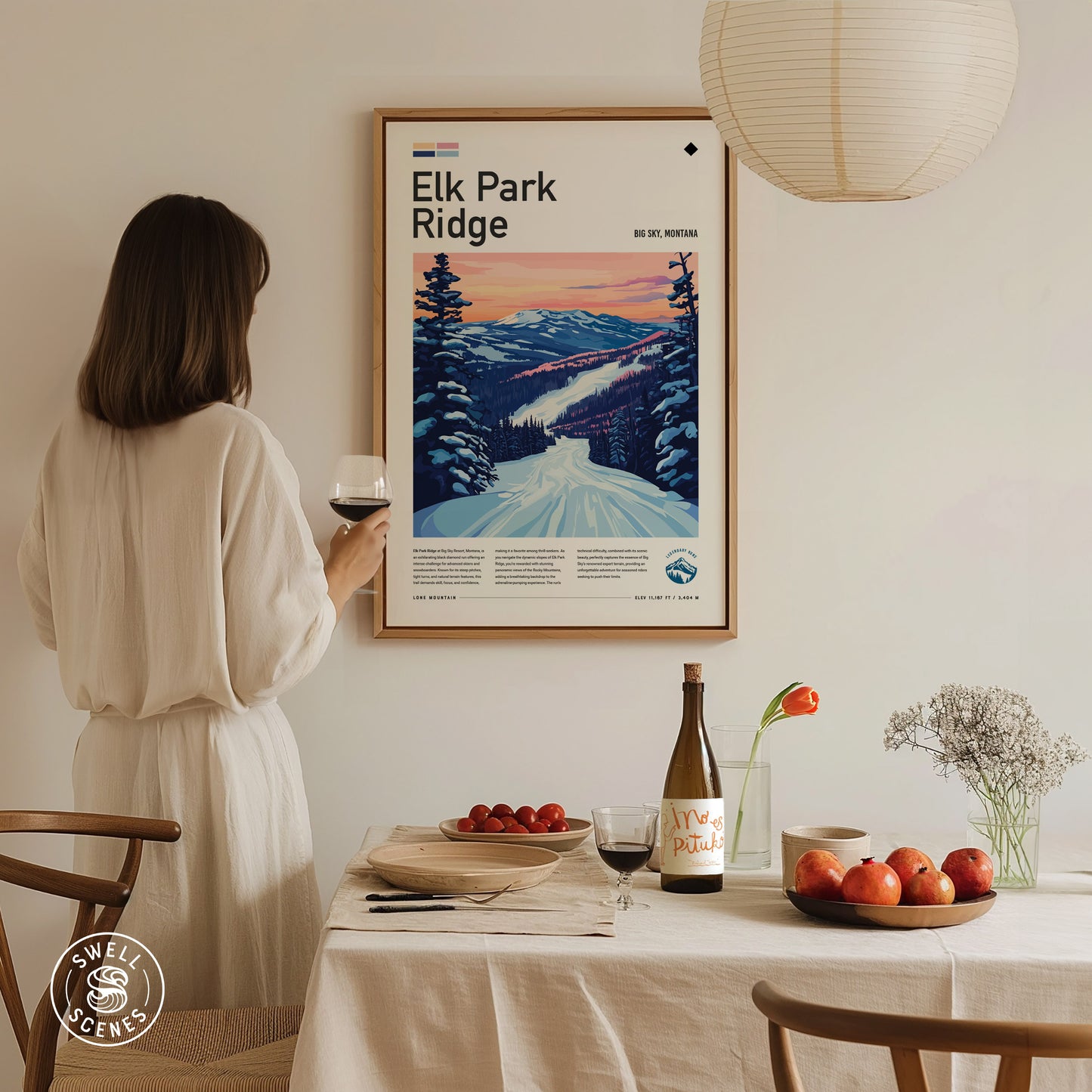 A woman in a white dress admires a framed Elk Park Ridge ski poster on a wall. A dining table is set with fruit, wine, and flowers.