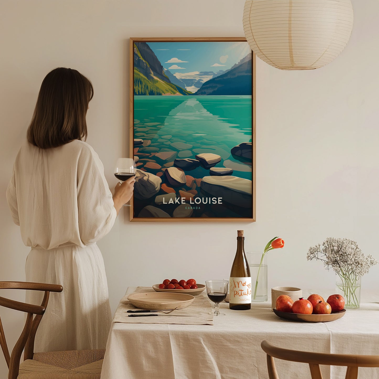 A woman in a white dress admires a Lake Louise poster on the wall while holding a wine glass. A table with fruit, wine, and flowers is in the foreground.