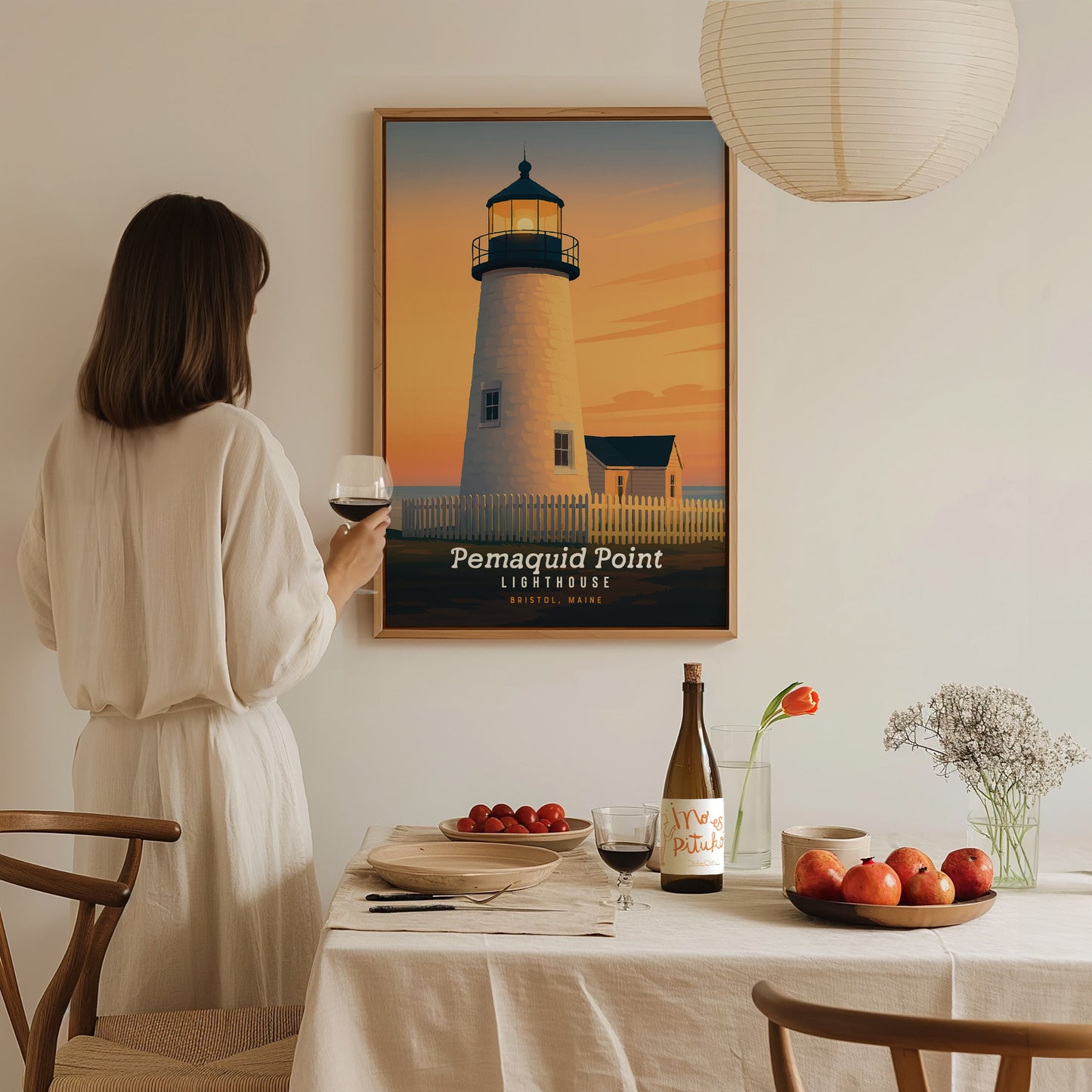A woman in a light-colored, flowing dress stands at a dining table, holding a glass of wine, while gazing at a framed poster of Pemaquid Point Lighthouse against a sunset backdrop.