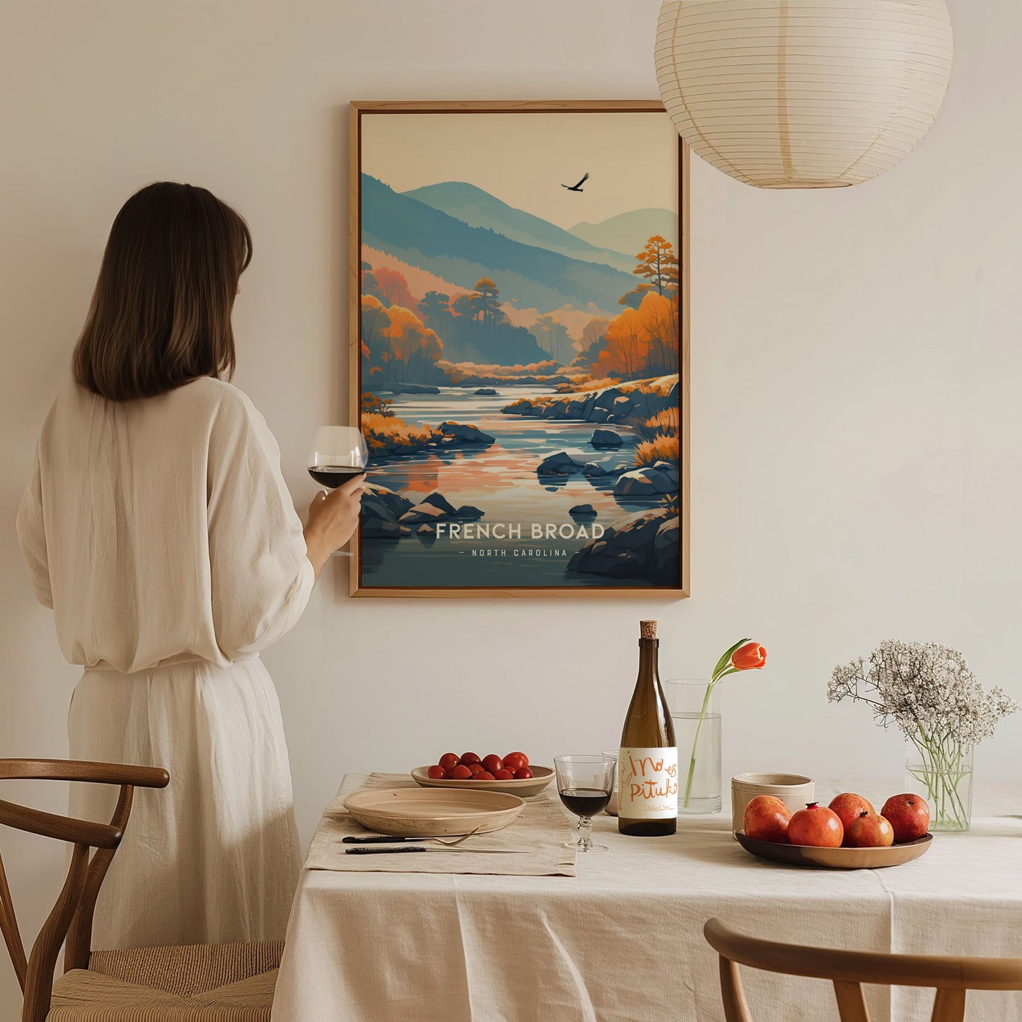 A woman in a white dress admires a framed French Broad River poster. A dining table with wine, fruit, and flowers is in the foreground, creating a serene atmosphere.