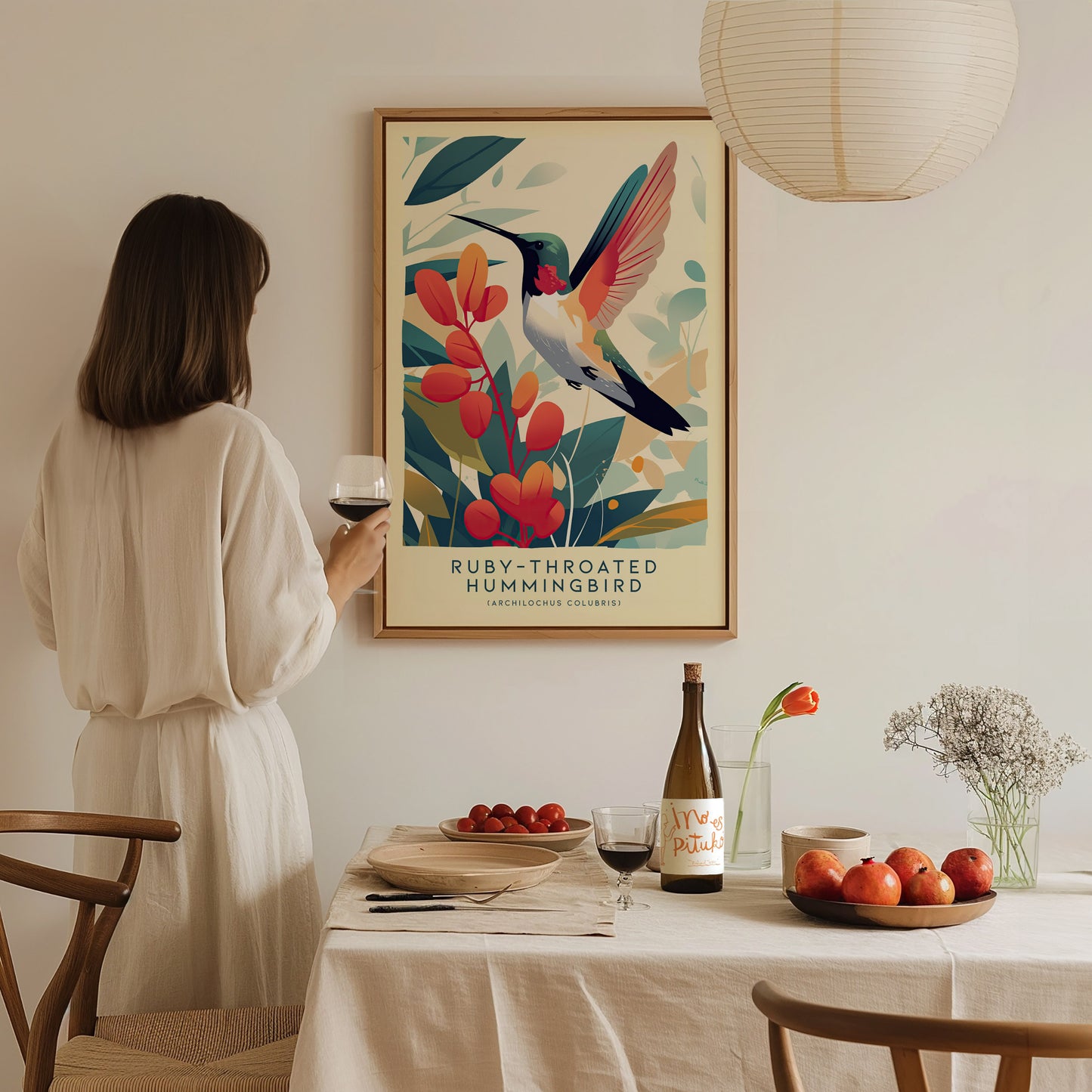 A person in a white dress admires a framed Ruby-Throated Hummingbird poster on a wall, with a dining table set with fruit and wine in the foreground.