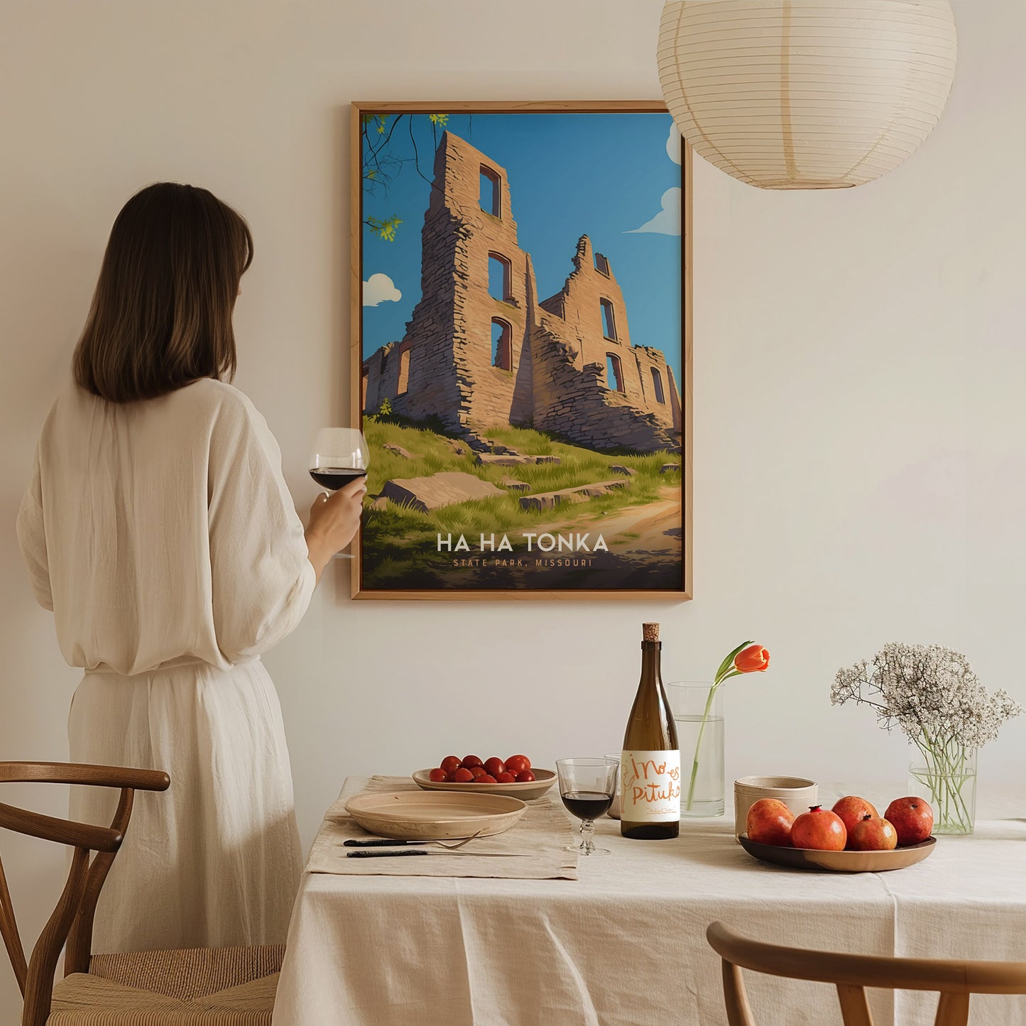 A woman in a white dress holds a glass of wine, admiring a framed Ha Ha Tonka State Park poster. A dining table with fruit, wine, and flowers is in.