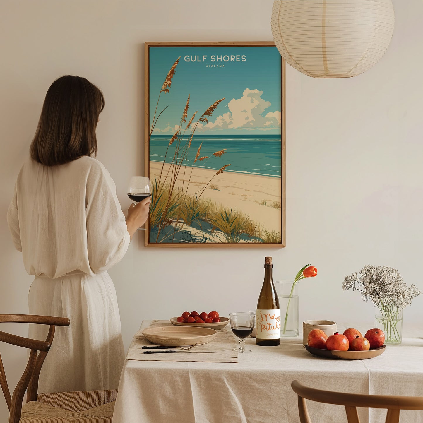 A woman in a white dress admires a Gulf Shores, Alabama beach poster on the wall, while standing beside a table set with fruit, wine, and a floral arrangement.