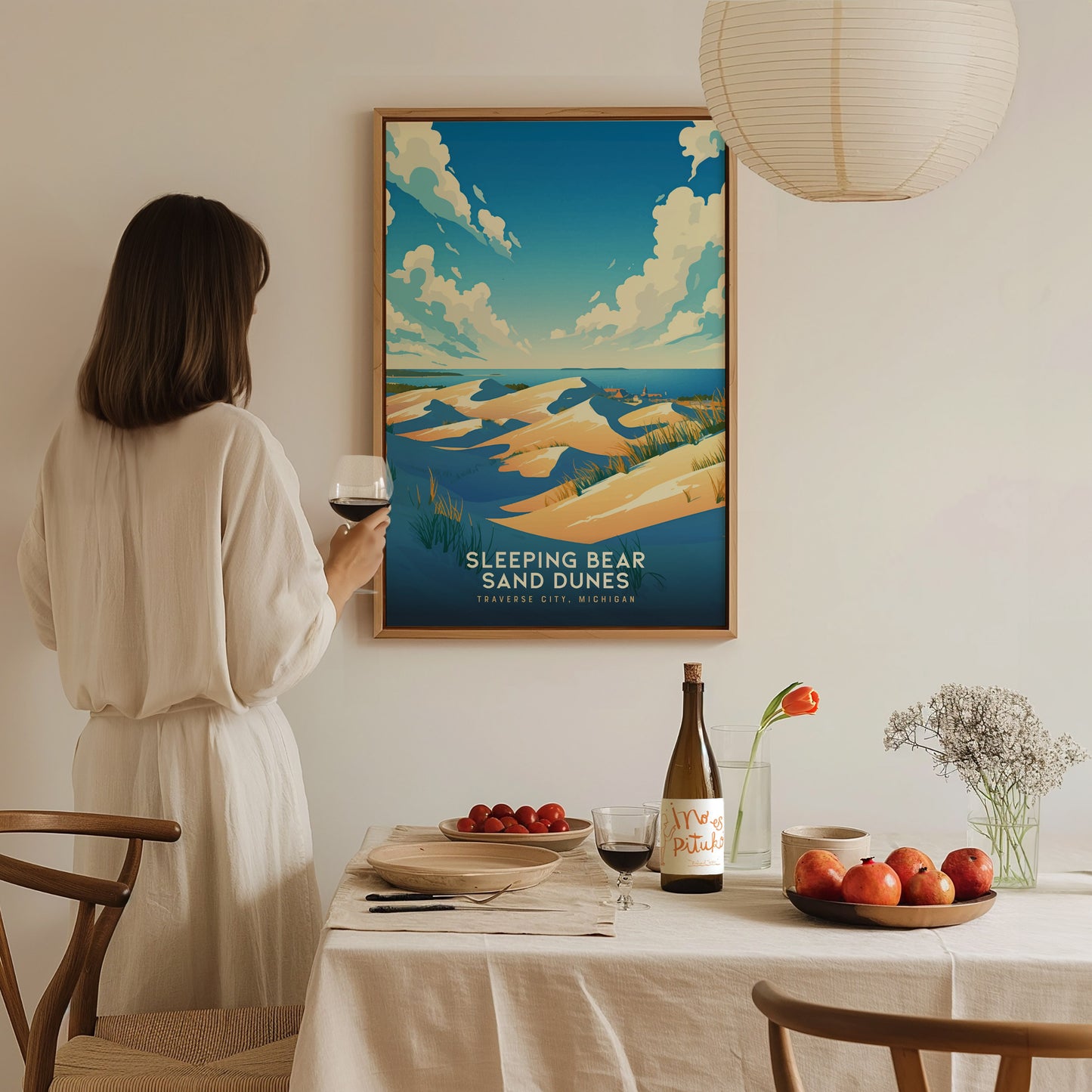 A woman in a white dress holds a glass of wine, admiring a framed Sleeping Bear Sand Dunes poster. The dining table is set with fruit, wine, and flowers.