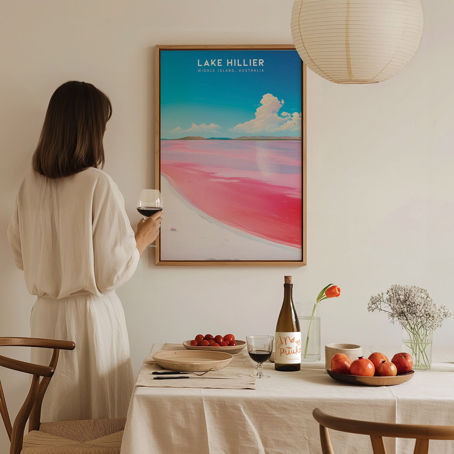 A woman in a white dress admires a framed Lake Hillier poster on the wall, with a dining table set with fruit, wine, and flowers in the foreground.