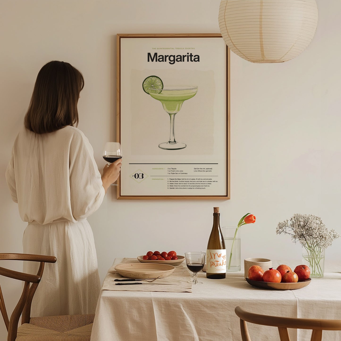 A woman in a white dress holds a wine glass while admiring a Margarita cocktail poster. The table is set with fruits, a bottle, and a vase with flowers.