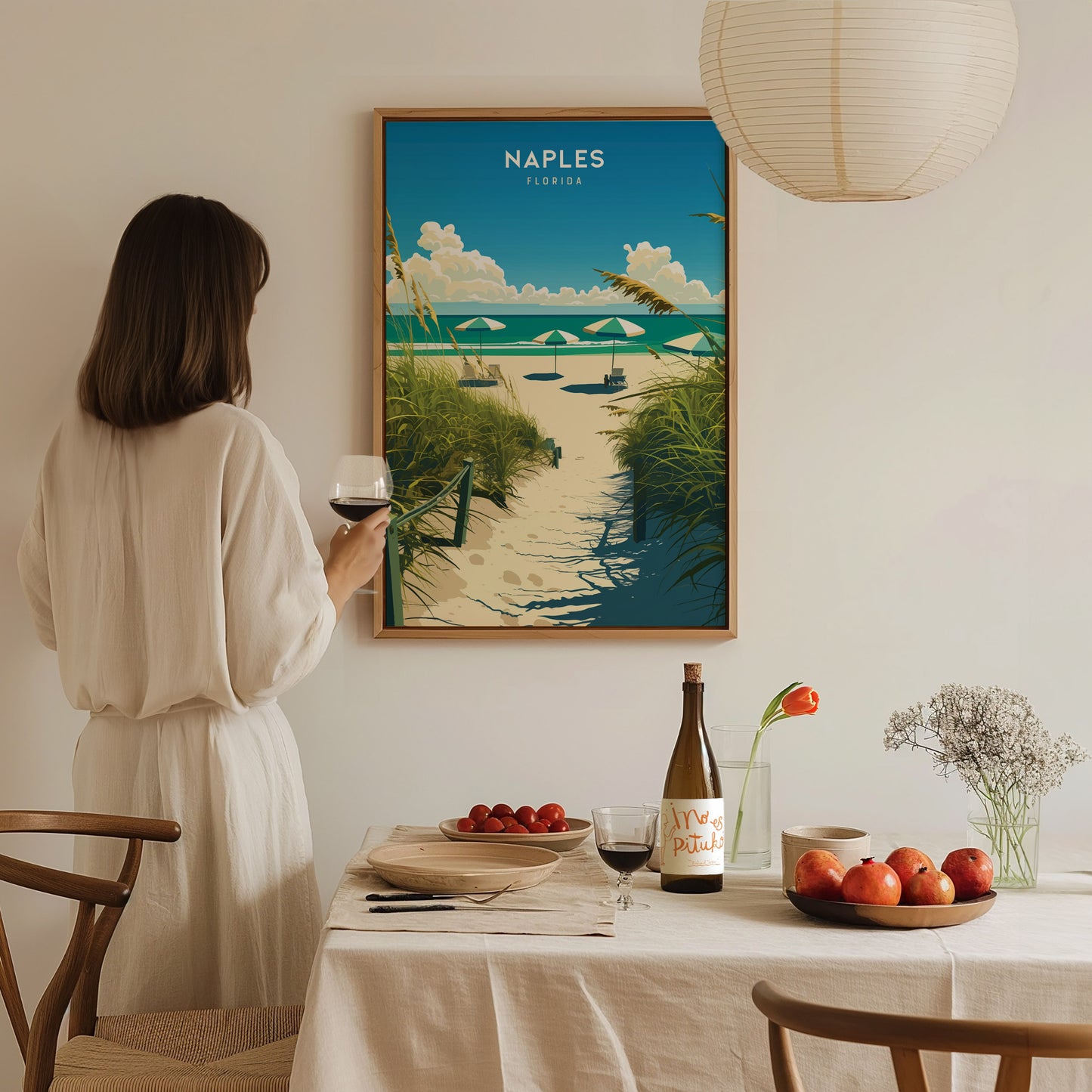 A woman in a white dress holds a glass of wine, admiring a Naples, Florida beach poster. The table is set with fruit, wine,.