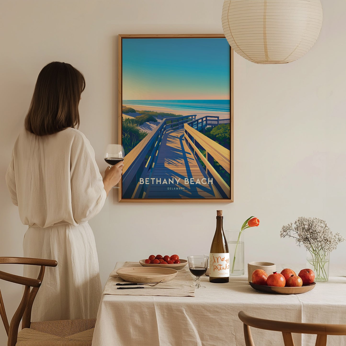 A woman in a beige dress admires a Bethany Beach poster on the wall, holding a glass of wine. A dining table with fruit and wine is in the foreground.